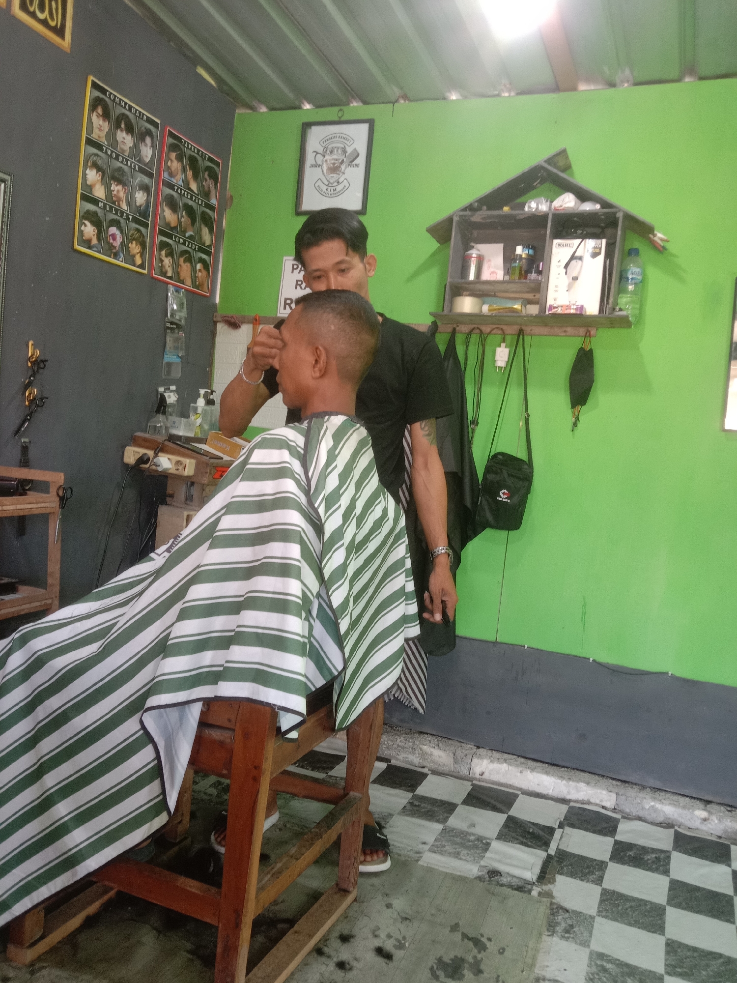 A barber is cutting the hair of a young man in a barbershop. The barbershop is decorated with pictures of different hairstyles on the wall, and there are shelves with various hair products and tools. The floor is tiled with a black and white checkered pattern. The barber is wearing a black t-shirt and the customer is wearing a striped cape. The barber is using scissors to cut the customer's hair.