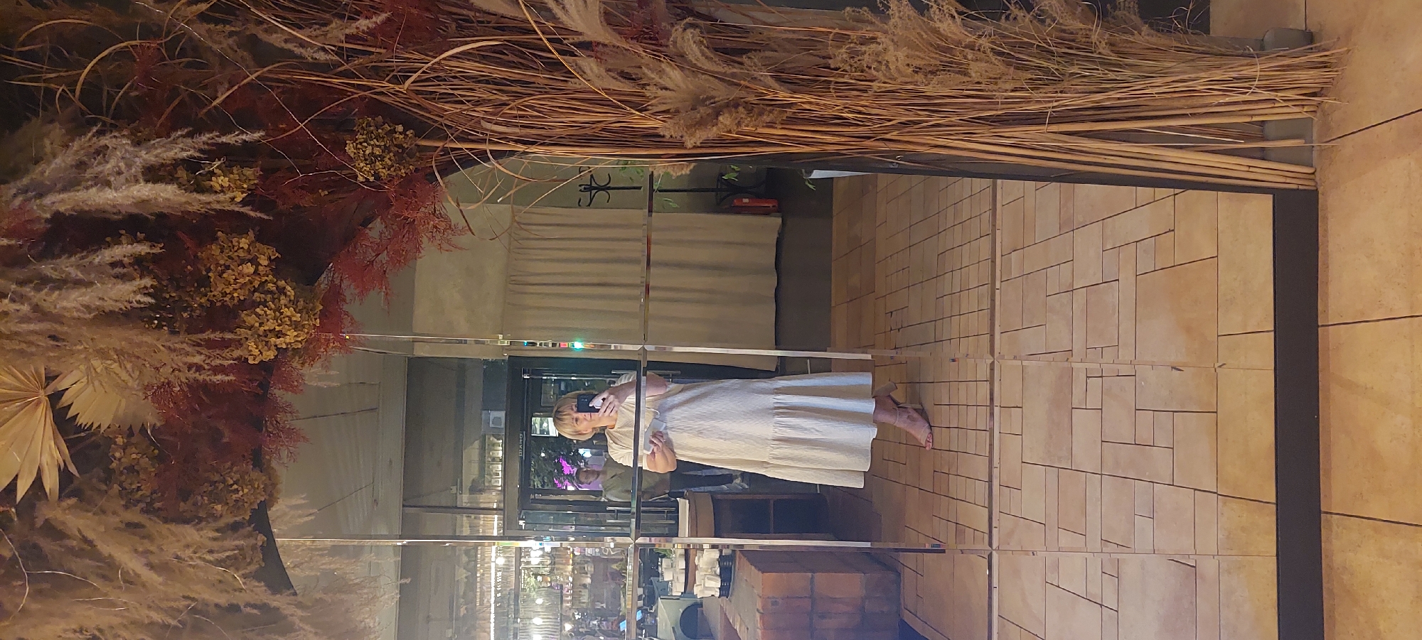 A woman is taking a mirror selfie in a cafe or restaurant in Kyiv, Ukraine. She is wearing a light-colored, striped dress and high-heeled sandals. The mirror is large and framed by a decorative arrangement of dried plants and grasses. In the reflection, a man can be seen standing in the background. The floor is tiled with a pattern of light brown and beige squares and rectangles. The environment appears to be a well-lit interior space with muted colors.