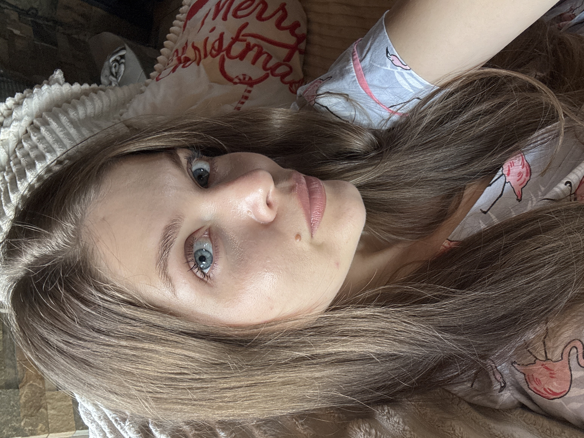 A close-up, waist-up photograph shows a young woman with long, light brown hair and striking blue eyes. She is looking directly at the camera with a neutral expression. Her skin appears clear with a slight sheen, and she has a mole on her cheek and a small blemish on her chin. She is wearing a grey pajama top with a pattern of pink flamingos. The background is softly blurred, showing a textured blanket and a pillow with red lettering that appears to say "Merry Christmas". The overall lighting suggests an indoor setting, possibly a bedroom.