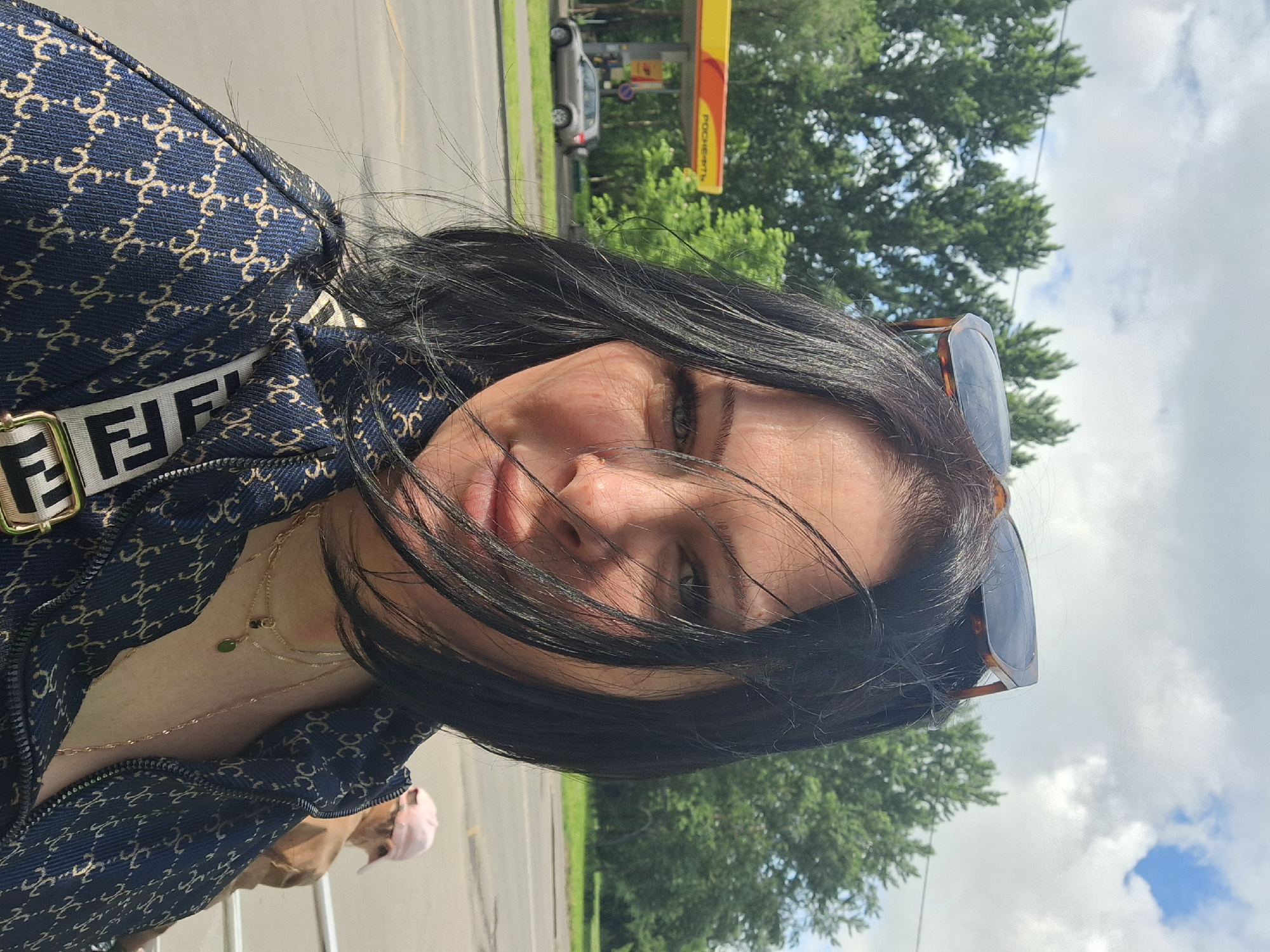 A woman with dark hair is shown in a selfie. She is wearing a blue jacket with a pattern and the words "FENDI" on the strap. She also has sunglasses perched on her head and a layered necklace. The background reveals an outdoor urban setting. Trees, a road, and a car are visible. A gas station with the name "POCHEON" is also visible. The gas station indicates the image was taken in Moscow, Russia.
