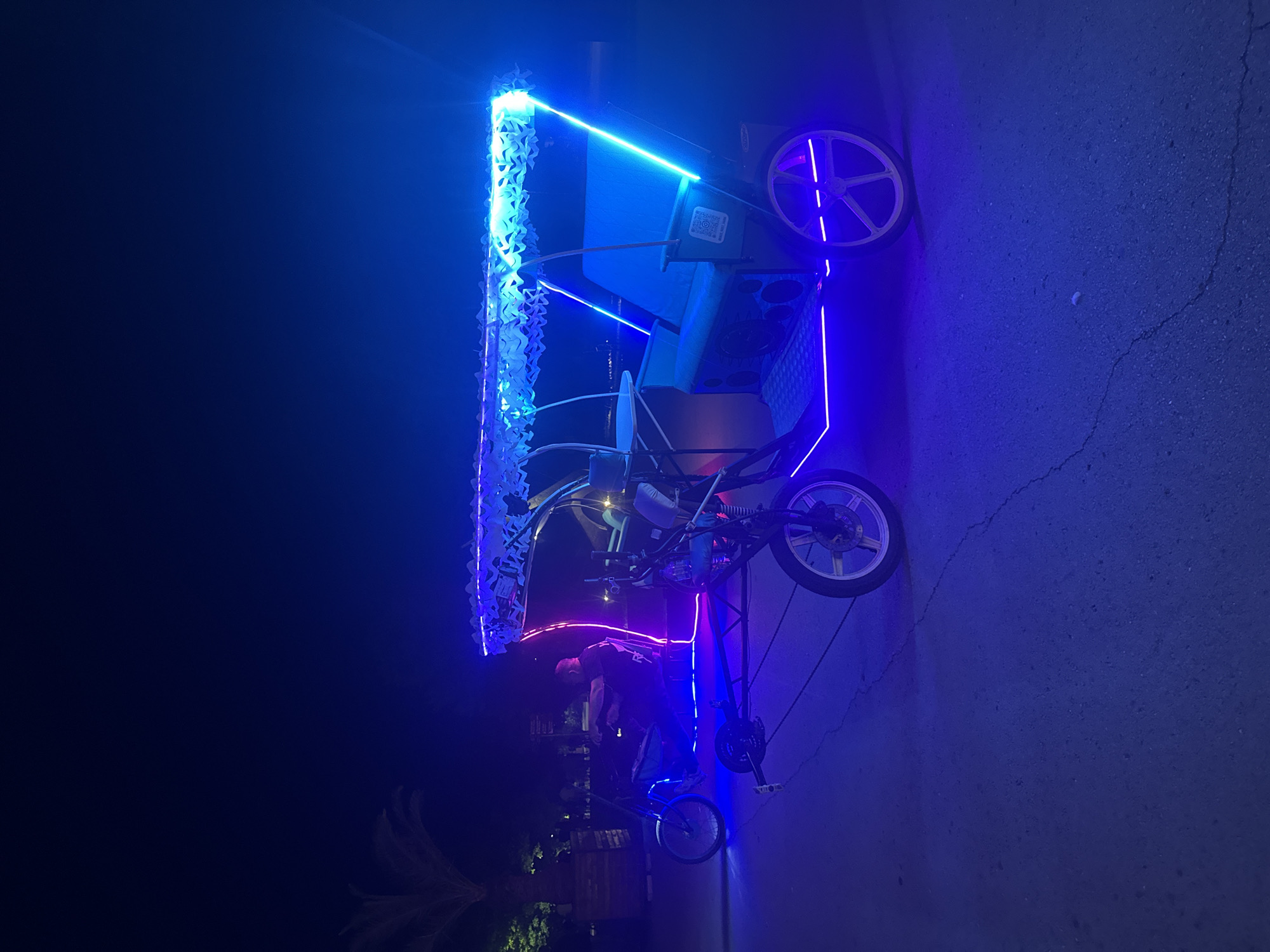 A low-angle, night-time shot captures a pedal-powered rickshaw, illuminated by vibrant blue and purple LED lights, parked on a cracked asphalt surface. The rickshaw is adorned with white, lacy streamers along its roof. Inside the rickshaw, what appear to be speakers are visible. A man, wearing a dark t-shirt and pants, is seen from the side, bending over and operating the vehicle. The scene is dark, with the only significant illumination coming from the rickshaw's lights, suggesting it's nighttime. The background is dark, with some faint hints of palm fronds and what might be building structures. The overall ambiance is festive and perhaps a bit unusual due to the bright, artificial lighting against the dark sky. There is a QR code visible on the side of the rickshaw.