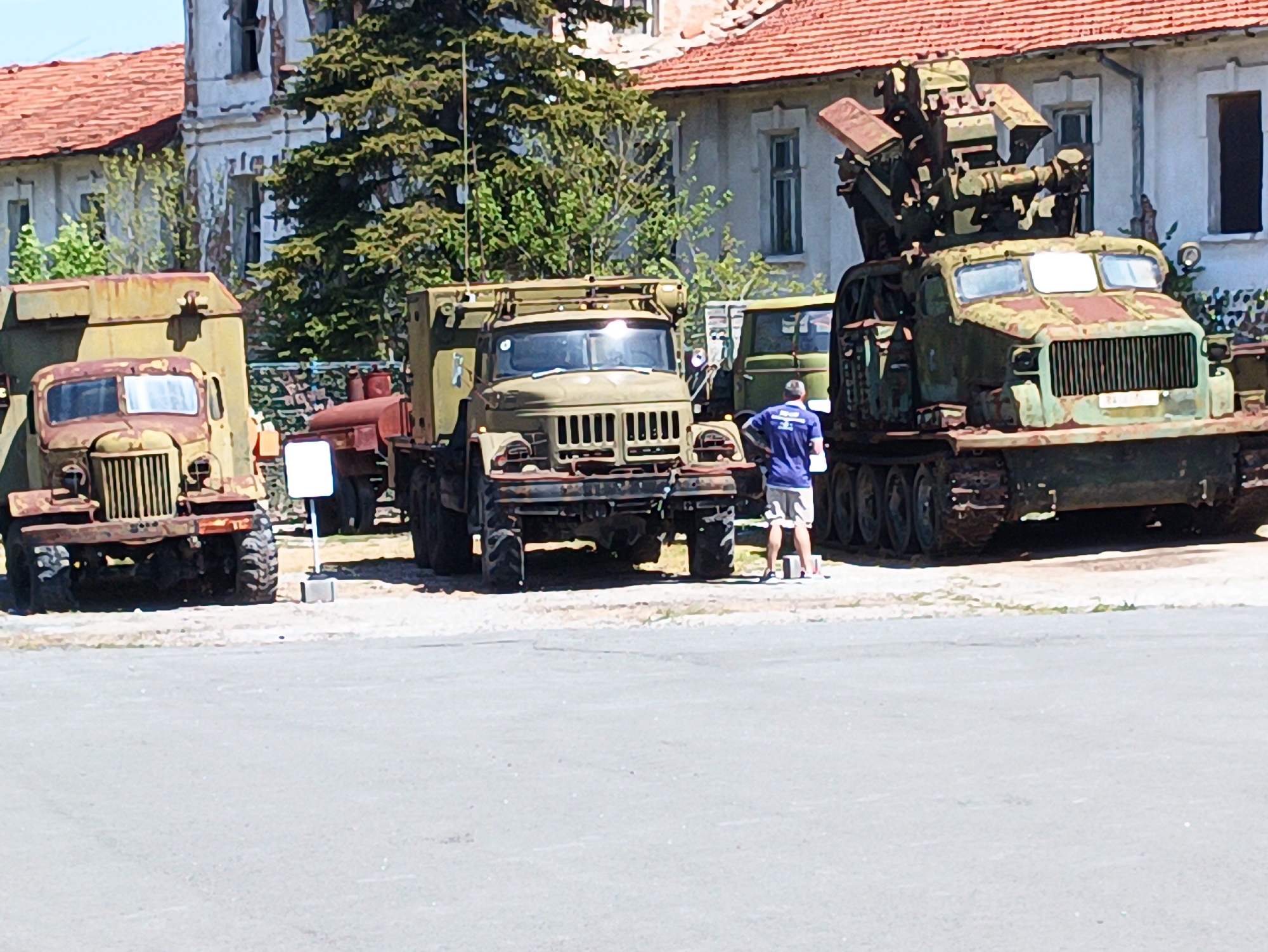 The image shows three old military vehicles parked in a lot, likely in Yambol, Bulgaria, based on visual characteristics of the architecture. The vehicles appear weathered and are likely on display. The closest vehicle to the right is a tracked military vehicle with a missile launching system on top. The middle vehicle is a cargo truck. To the left is another cargo truck. A man in a blue shirt and shorts is standing near the center vehicle, seemingly examining it. The backdrop consists of a large building with a red tiled roof and several windows and doors. The surrounding environment includes trees and an asphalt surface.