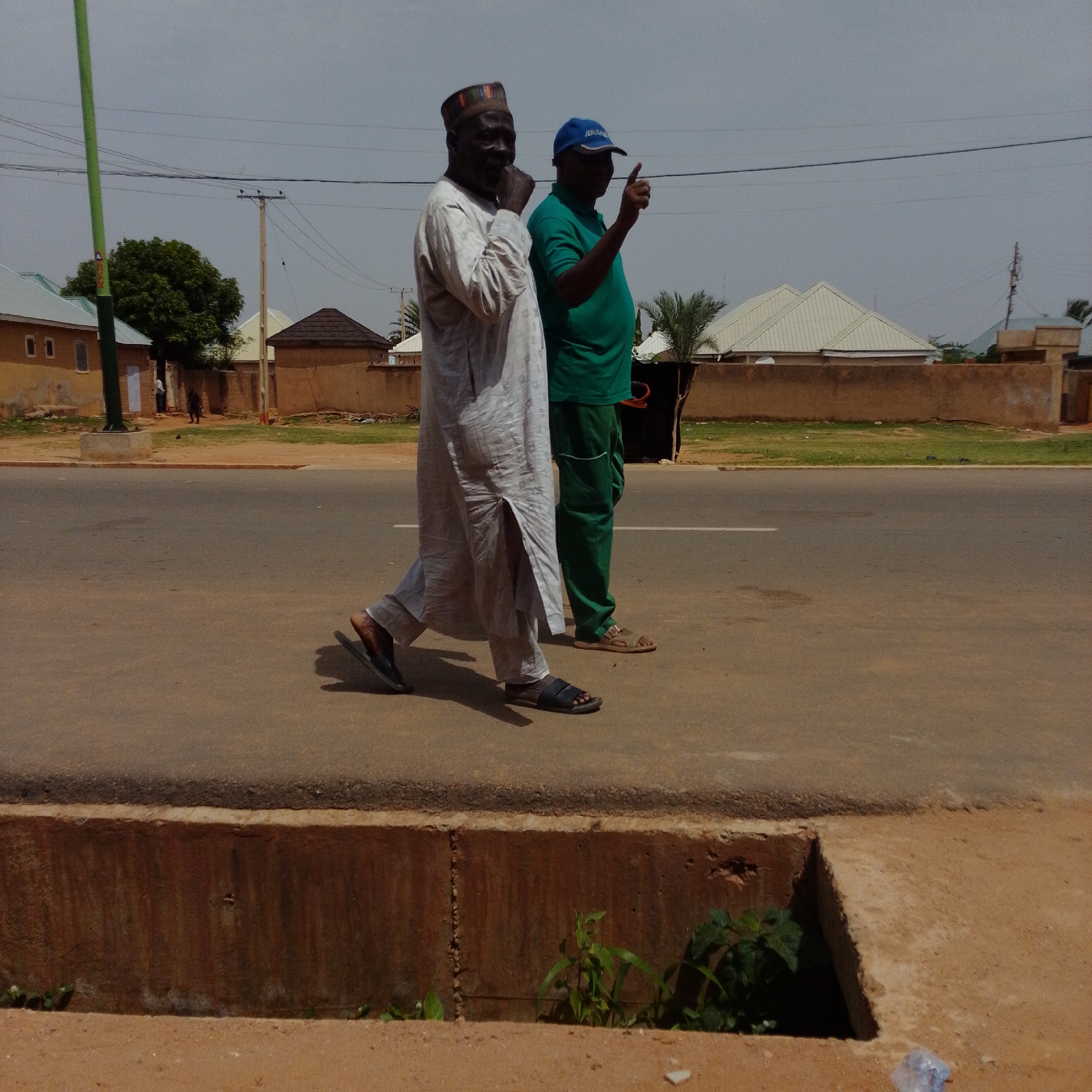 This is a medium shot, outdoor photograph taken on a cloudy day in Gombe, Nigeria. The foreground features a partial view of a drainage ditch with a concrete structure, some sparse green vegetation growing out of it, and sandy ground. The midground shows a paved road with a white line marking. Two men are walking on the road. The man on the left is wearing a light-colored, long robe with a patterned cap and dark sandals. He is walking with his body angled slightly to his left. The man on the right is wearing a green t-shirt, green pants, and a blue baseball cap. He is walking with his body angled slightly to his right and is pointing his right index finger upwards. In the background, there are several buildings with corrugated metal roofs, some trees, and utility poles with power lines. A green pole with a concrete base is visible on the far left. The overall scene depicts a street in a residential or commercial area.