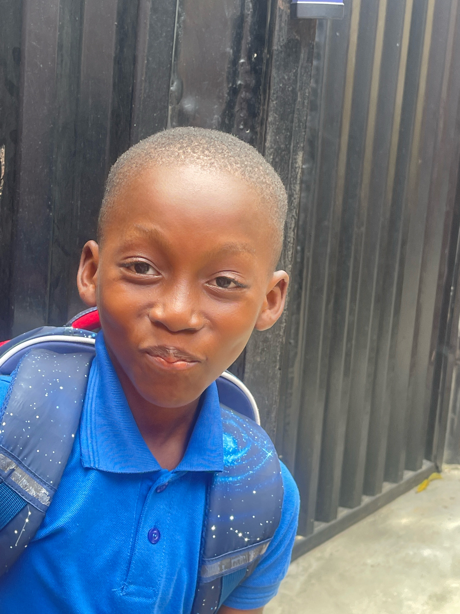 A young boy wearing a blue collared shirt and a blue backpack with a galaxy print stands in front of a dark, corrugated metal fence. The boy has short, dark hair and is looking towards the camera with a slight smile. His backpack has white straps and a reflective strip. The fence appears to be part of a building's exterior. The ground in front of the fence is concrete, and there is a yellow leaf on the ground to the right of the boy. The lighting suggests it is daytime, possibly outdoors. The background is out of focus, highlighting the boy as the main subject.