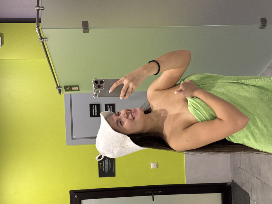 An indoor scene in a fitness or spa facility in Vniissok, Russia, features a young adult female captured in a mirror selfie. The female subject has long, dark hair and is wearing a white felt sauna hat and a light green towel wrapped around her body, covering her from just above her breasts to her upper thighs. She holds a grey smartphone with a stylized case in her right hand, angling it to photograph her reflection. She is smiling broadly, with her head slightly tilted, and looking towards her left. Her left hand is subtly placed over the towel near her chest. The background consists of a vibrant lime-green painted wall. To the left, a dark-framed door is partially visible. To the right, there are light green frosted glass partitions, likely for shower stalls or changing rooms, with a horizontal metal bar across the top. On the lime-green wall, a black rectangular sign with white Cyrillic text is visible in reflection, stating "СЛЕДУЙТЕ ПРАВИЛАМ" (FOLLOW THE RULES). Below it, another sign's reflected text partially reads "ОБЯЗАТЕЛЬНОЕ СОБЛЮДЕНИЕ" (MANDATORY COMPLIANCE). The floor is tiled in a light grey color.
