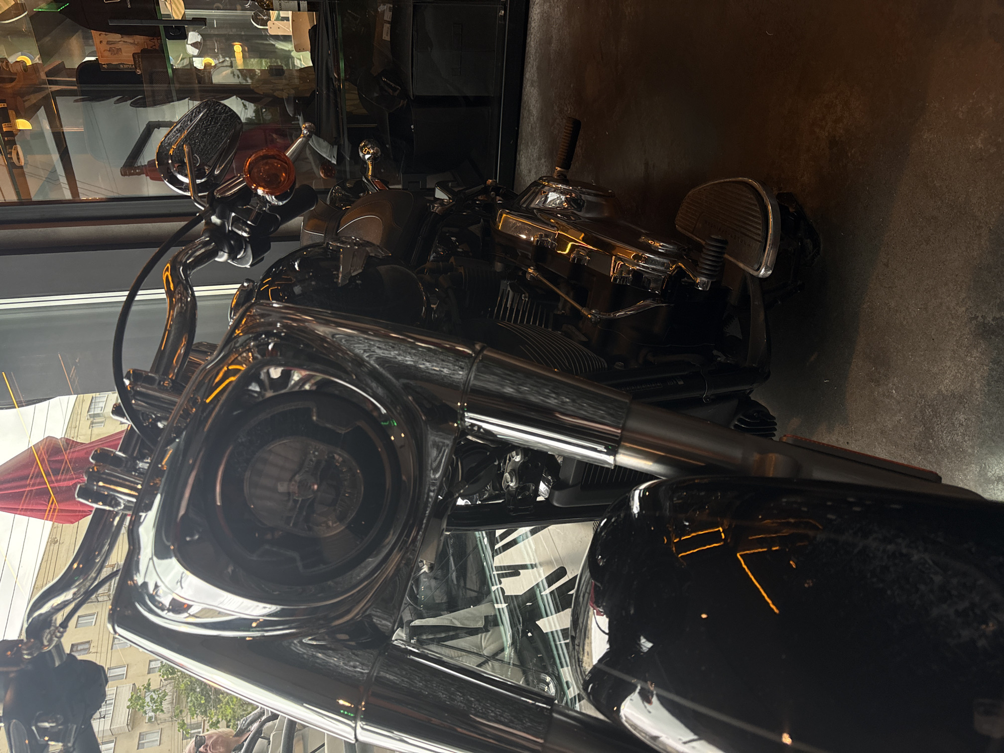 A low-angle, close-up photograph displays the front and right side of a stationary, dark-colored, heavily chromed cruiser-style motorcycle, positioned indoors within a commercial establishment in Izhevsk, Russia. The motorcycle features a prominent round LED headlight, polished chrome forks, handlebars, and visible engine components. An amber turn signal is present on the right handlebar assembly. A wide, textured footpeg is situated on the right side of the motorcycle.
The motorcycle rests on a dark, unpolished concrete floor. A large glass window to the left reflects an exterior urban environment, showing the upper portions of light-colored, multi-story buildings with visible windows and balconies, along with green foliage and an overcast sky. A large red object, likely an umbrella or awning, is partially reflected near the top center. A reflection of a person with sunglasses and light hair is visible in the window near the motorcycle's front fender. To the right, behind the motorcycle, glass display shelves are visible, containing various items, including a bottle of Baileys, a product with Cyrillic script, and another item labeled "DIMONA." Artificial yellowish light sources, both visible and reflected, illuminate the interior and highlight the motorcycle's chrome surfaces.