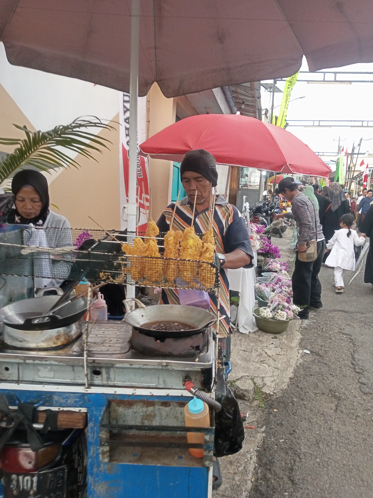 In Pandansari, Indonesia, a street vendor stands behind a cart, frying food items on sticks, possibly corn dogs or similar snacks, in a large wok filled with hot oil. He is wearing a dark beanie and a striped shirt. To his left, another vendor, a woman wearing a hijab, is also preparing food. In the background, a busy street scene unfolds with other vendors, customers, and motorcycles. A red umbrella shades some of the stalls, and flowers are displayed for sale. The overall impression is of a lively, outdoor market.
