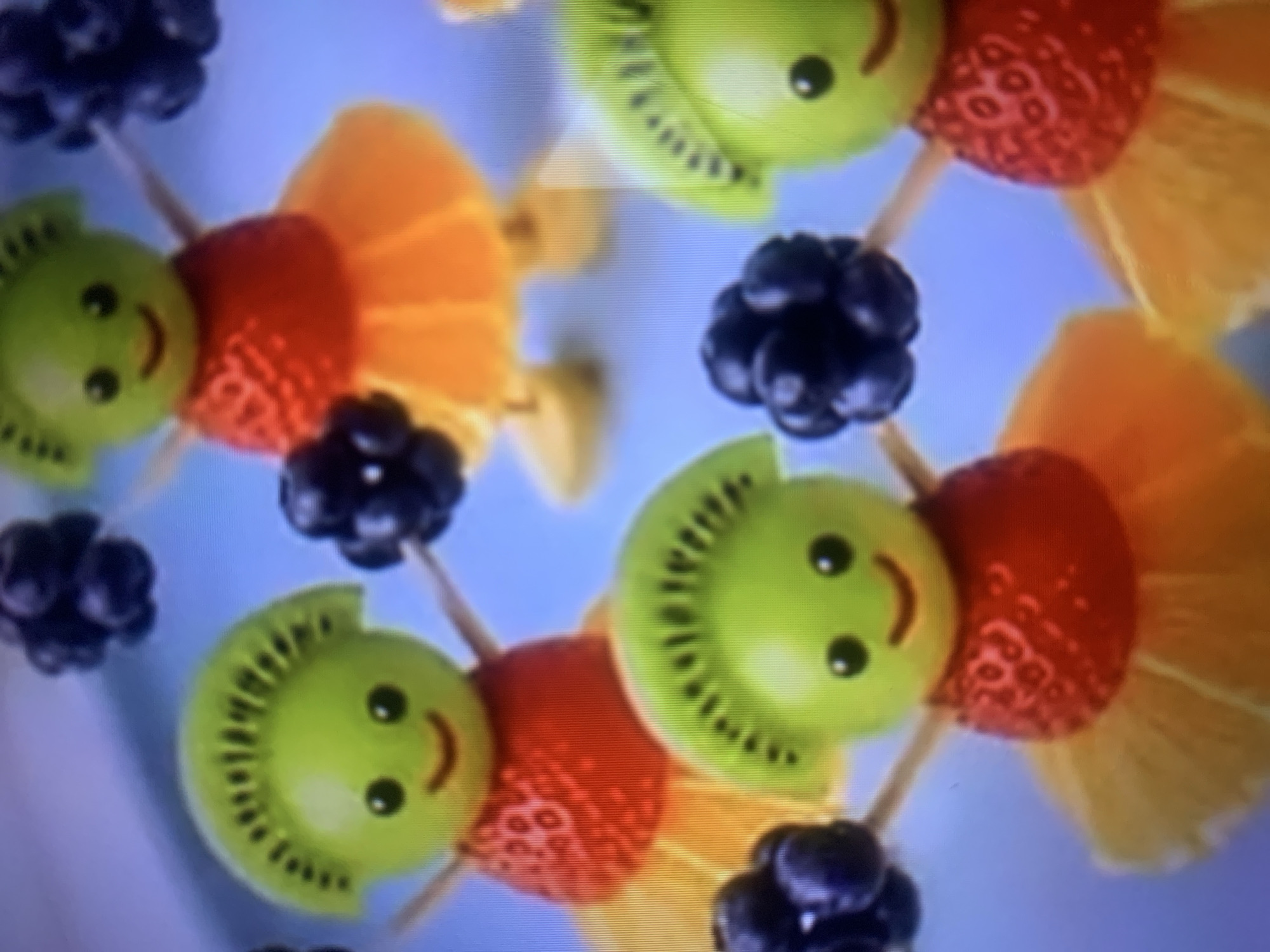 This is a close-up, eye-level, medium shot of several fruit figures arranged on a light blue surface, possibly a tablecloth or board. The figures are crafted to resemble cartoon characters, with bodies made of strawberries and heads made of green grapes topped with slices of kiwi fruit. Their eyes are black dots, and their mouths are small curved lines, giving them cheerful expressions. Each figure is adorned with a skirt made of thinly sliced orange or yellow fruit, and they have blackberries for hands. The lighting is bright and even, suggesting an indoor setting or possibly an outdoor setting with diffused light. The focus is sharp on the foreground figures, with a slight softening of detail in the background figures. There are no discernible architectural elements or contextual clues that would place this scene in Almaty, Kazakhstan, or any specific city. The overall impression is of a whimsical and playful creation, likely intended for a children's party or a decorative food display.