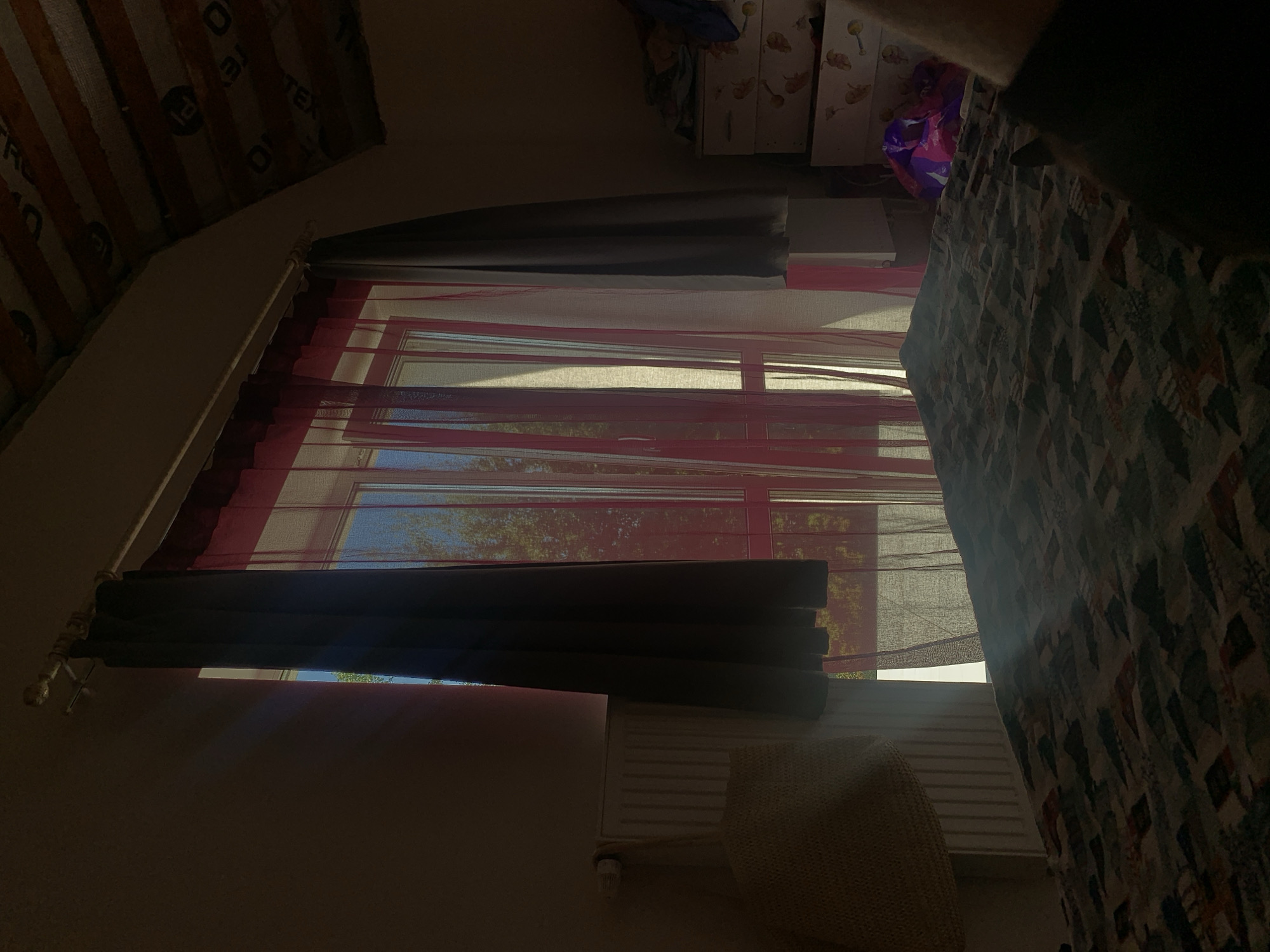 The image depicts an interior scene, likely a bedroom, with a large window covered by two layers of curtains: a dark outer curtain and a sheer pink inner curtain. Sunlight streams through the window, casting diagonal beams of light across the room and onto a bed with a patterned comforter. A radiator is visible beneath the window, with a woven bag hanging on it. To the right of the window, there is a dresser with drawers decorated with a fruit motif. A brightly colored plastic bag is on top of the dresser. The ceiling appears to be sloped and has exposed beams. The scene is dimly lit, with the sunlight providing the primary illumination. The image quality is somewhat grainy, suggesting it was taken in low light conditions. No people are visible in the image. The scene does not provide any specific visual cues to pinpoint its location in Minsk, Belarus.