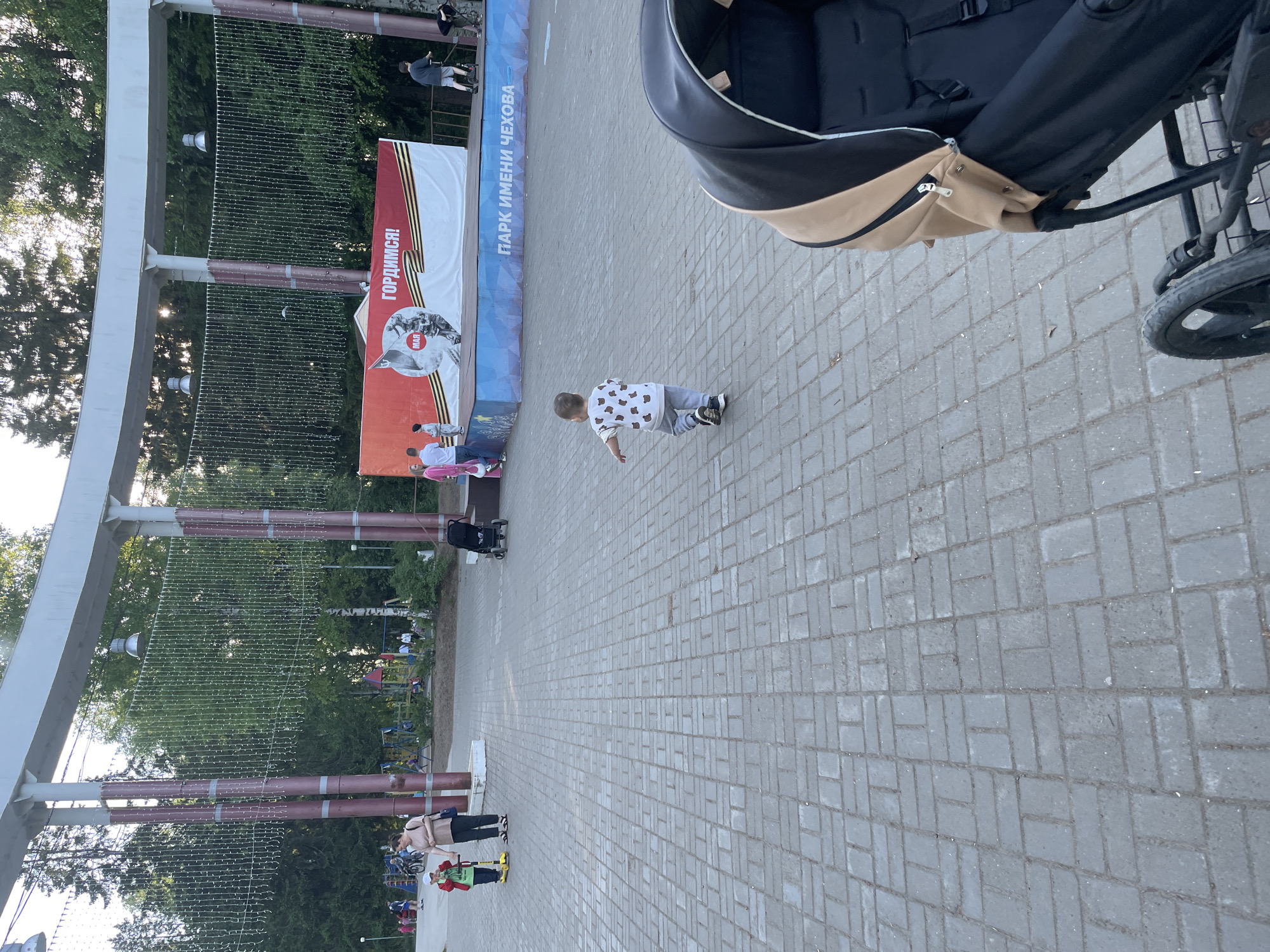 A photo taken outdoors at a park in Perm, Russia, shows a child crawling on a brick-paved walkway. The child is wearing a white shirt with black bear-like graphics and gray pants. To the left, a large sign is visible with Russian text, and other people are standing near the sign. A stroller is partially visible in the upper right corner. The scene suggests a public park or recreational area, possibly named after Chekhov, based on the sign, with greenery in the background. String lights are hung from a large structure in the park.