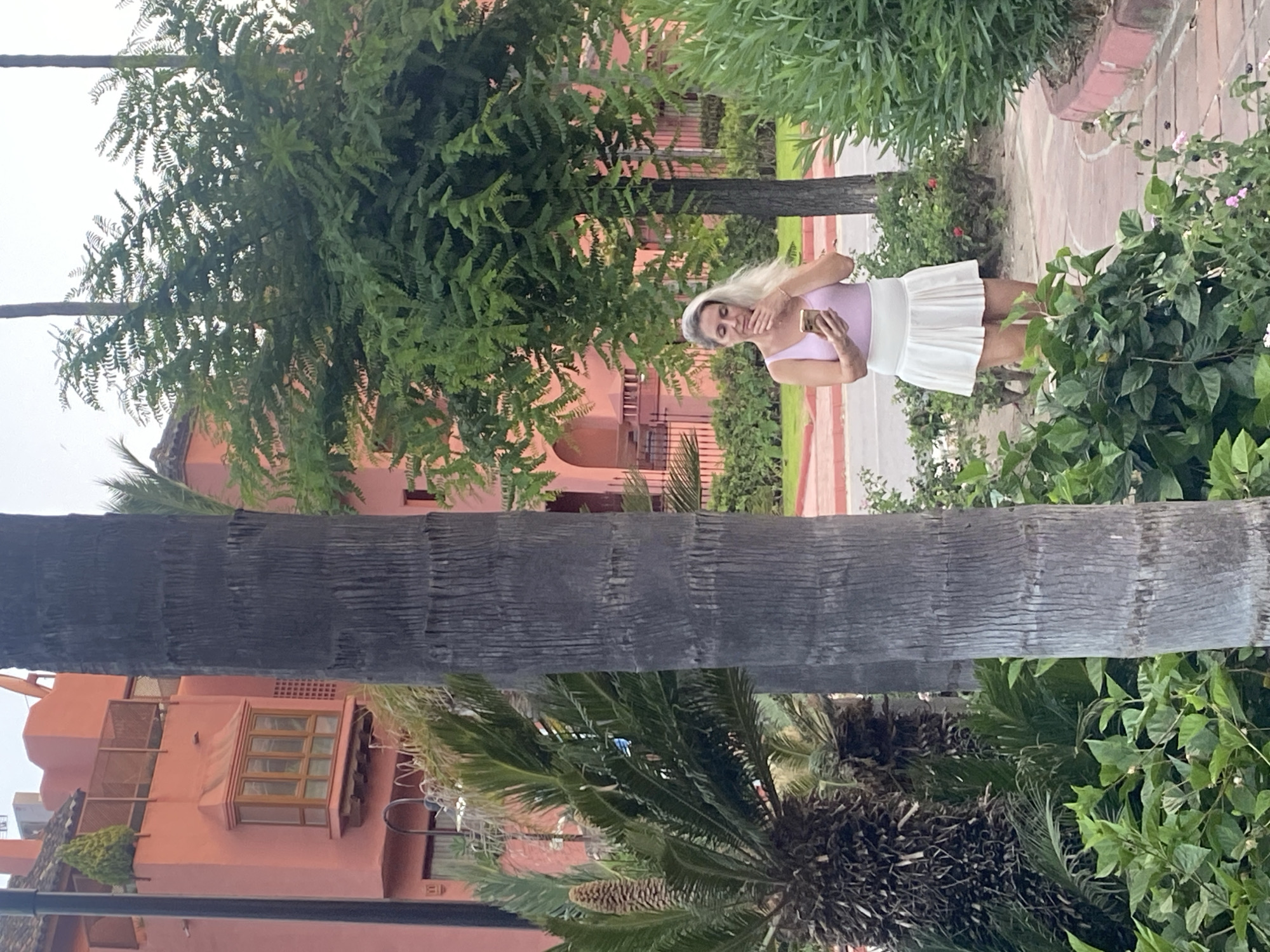 A woman with blonde hair stands in a garden, holding a smartphone in her hands as if taking a selfie. She is wearing a light purple tank top and a white pleated skirt. The woman is looking down at her phone, and her hands are raised to her face, obscuring part of her expression. She appears to be in a relaxed pose, possibly enjoying a moment of solitude in the lush greenery.
The background features a terracotta-colored building with balconies and windows, suggesting a residential or resort-like setting. The building is surrounded by a variety of plants, including trees with dense foliage and palm trees with rough bark. A paved pathway is visible to the right of the woman, bordered by more greenery and some small pink flowers. The lighting suggests it is daytime, possibly overcast. The overall impression is one of a pleasant, outdoor environment, likely in a warm climate given the vegetation. The scene seems to capture a casual, everyday moment, with the woman engrossed in her device amidst the natural beauty.