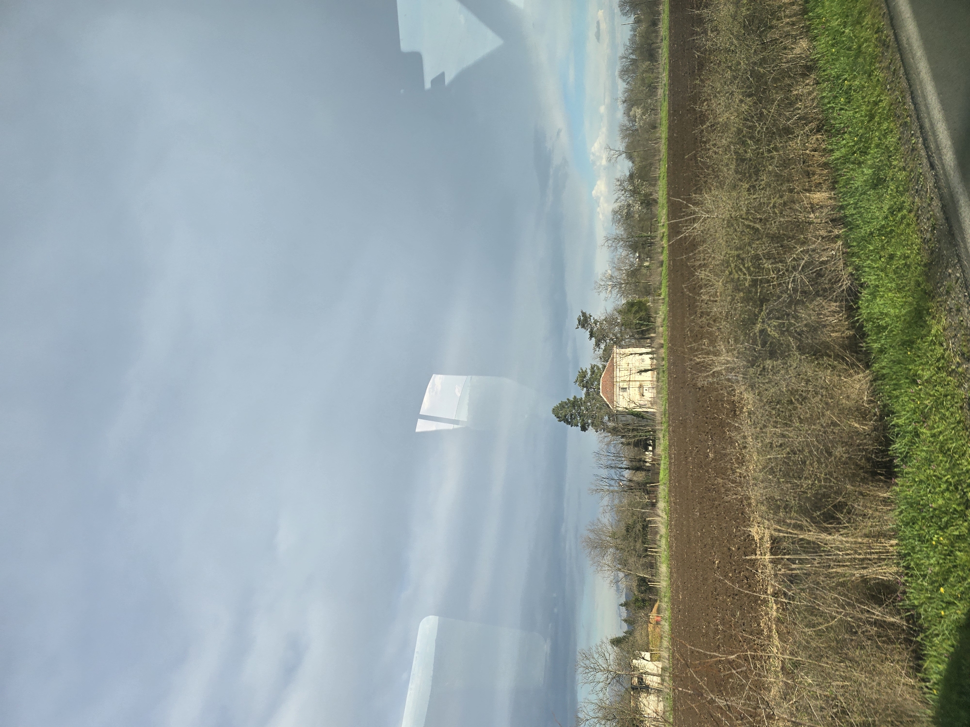 The image shows a landscape viewed through a window, with reflections obscuring parts of the scene. The landscape includes a large field of plowed earth, a strip of green grass along the edge of a road, and scattered trees. In the distance is a light-colored building with a red roof, possibly a house or small chateau. The sky is overcast with patches of blue visible, suggesting a cloudy day. The image is oriented in an unconventional vertical format. There is no indication of the city of Kabile, Bulgaria. No people are visible.