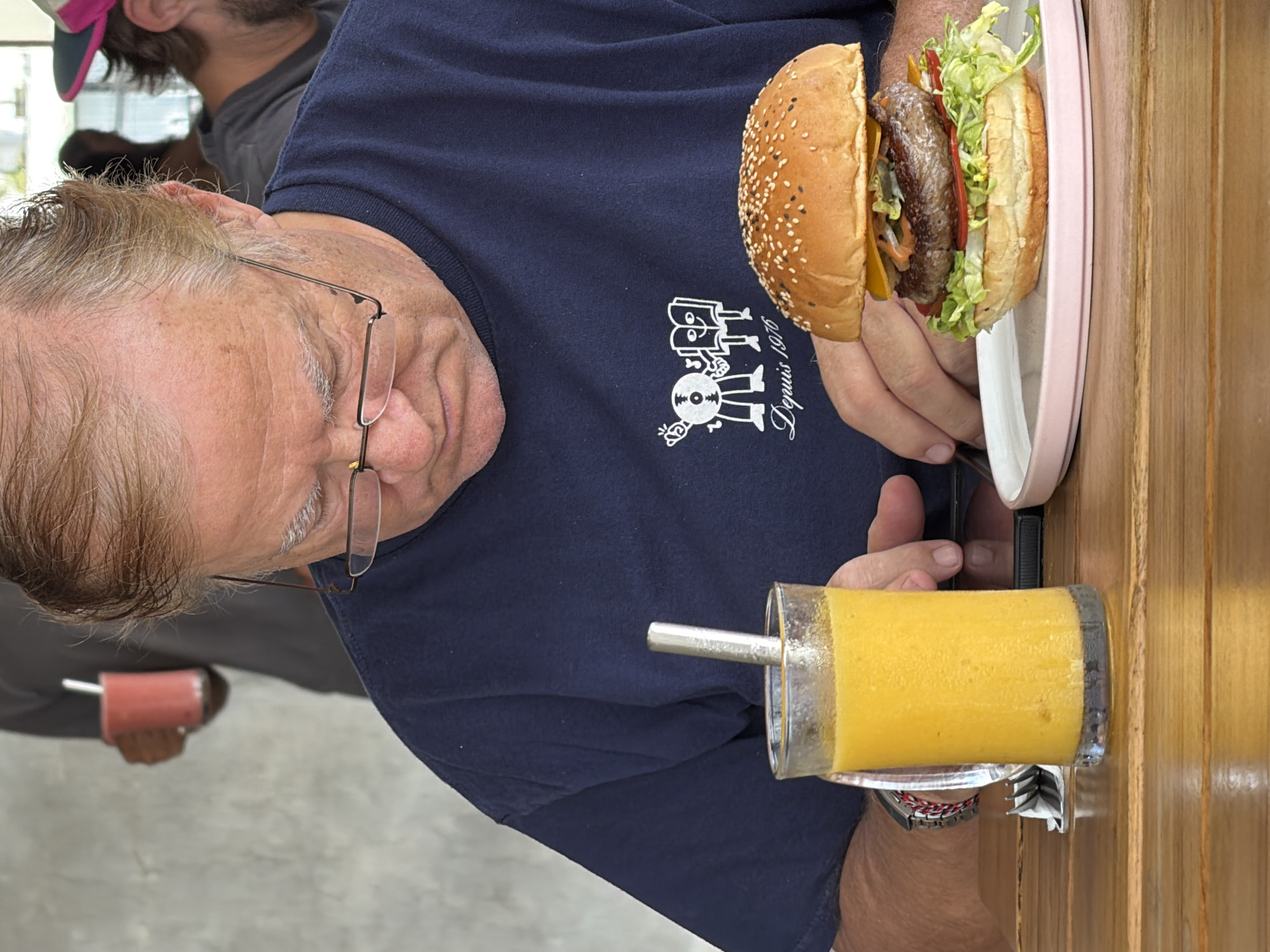A close-up, medium shot shows a middle-aged Caucasian man wearing glasses and a dark blue t-shirt sitting at a wooden table. He is looking down at his phone, which is partially visible on the table. In front of him, a glass of orange juice with a metal straw stands to the left, and a burger on a pink plate sits to the right. The burger is topped with sesame seeds, lettuce, tomato, and cheese. The man's t-shirt features a graphic with two cartoon characters and the text "Depuis 1976." The background is blurred, showing glimpses of other patrons, one of whom is holding a pink drink. The setting appears to be a casual eatery in Mengwi, Indonesia, as indicated by the context provided.