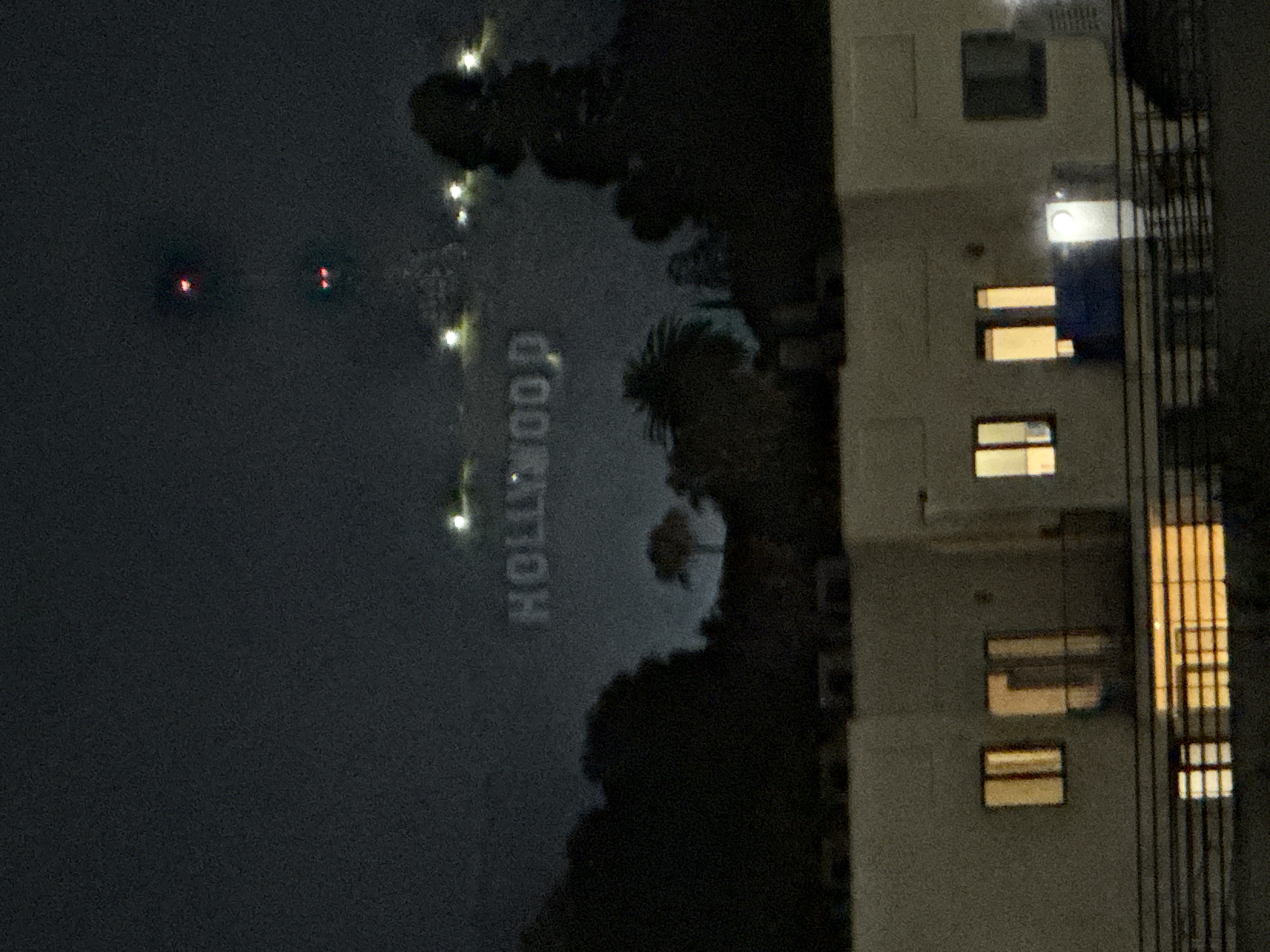 The image depicts the Hollywood sign illuminated at night, viewed from a high vantage point in Los Angeles, California, United States. The sign is partially obscured by trees. A tall building with multiple lit windows is visible to the right. Distant red lights are seen further away, suggesting a high-rise building or transmission tower. The sky appears dark.