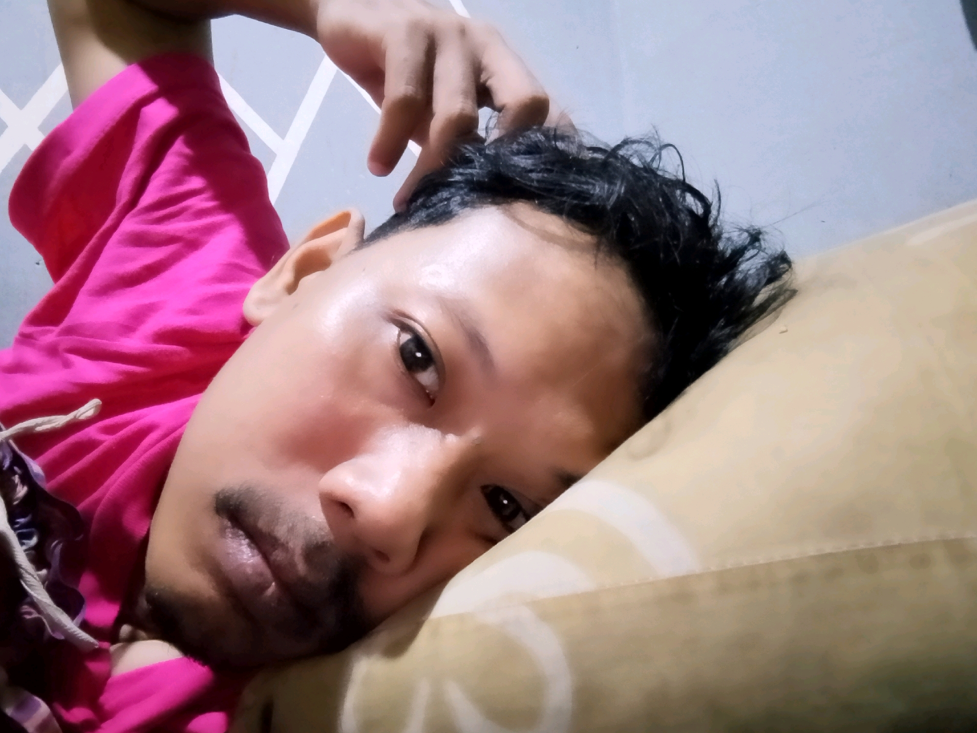 A close-up, low-angle selfie shows a person lying down on their side, propped on a pillow, and looking directly at the camera. The individual has dark, messy hair and is wearing a bright pink t-shirt. Their right hand is raised and resting on their forehead, fingers gently touching their hair. The background consists of a light blue wall with a geometric pattern and a beige pillow. The image appears to have been taken indoors, possibly in a bedroom, in a residential setting within Ciomas, Indonesia. The lighting suggests it is daytime.