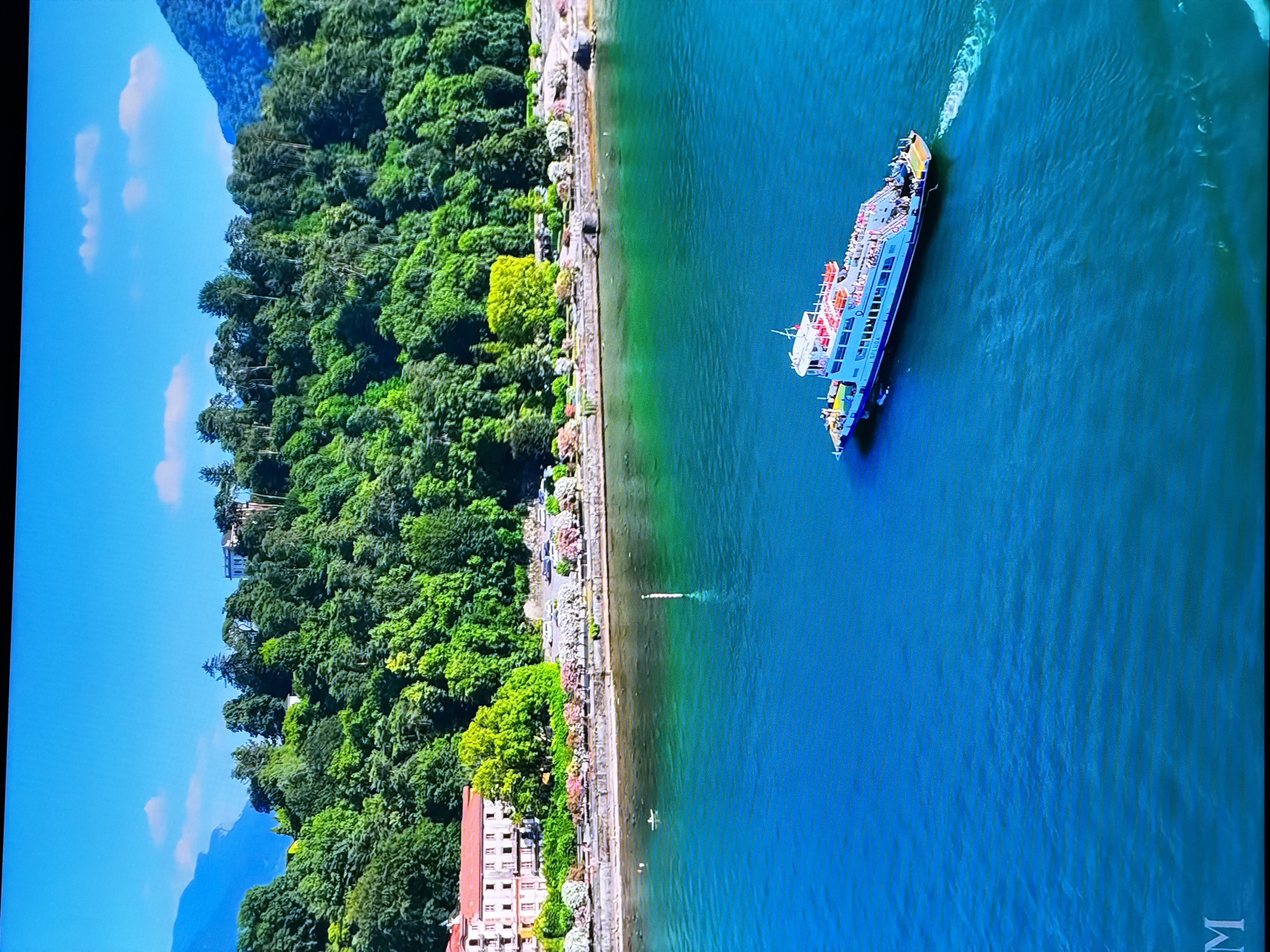 The image shows a scenic view of a body of water with a ferry boat sailing on it. In the background, there is a lush green hillside covered with trees, and a large building with a red roof is visible on the slope. The sky is bright blue with a few white clouds. The water is a deep blue color, transitioning to a turquoise hue closer to the shore. The ferry boat is white with blue trim and appears to be carrying passengers on its decks. The ferry is creating a wake as it moves through the water. There is a small white object, possibly a buoy or marker, in the water near the shore. The shoreline is lined with trees and some decorative landscaping. The overall impression is a serene and picturesque landscape. There is no discernible connection to the city of Chirchik, Uzbekistan.