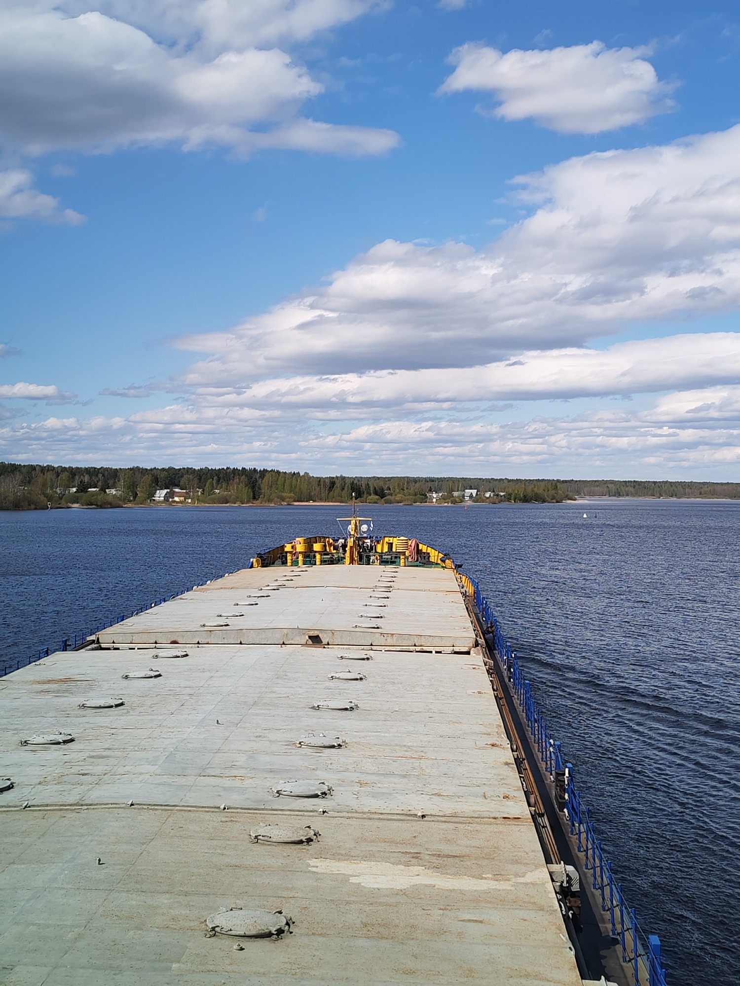 A long, flat-topped barge is centered, cutting through a body of water, with a view from the stern towards the bow. The barge is gray with a series of oval hatches along its surface and a blue railing along its right side. At the bow, there are yellow and black structures. The water is dark blue with gentle ripples. Across the water is a line of trees and some visible structures, suggesting a shoreline. The sky is blue with scattered white clouds. The location is Irdomatka, Russia.