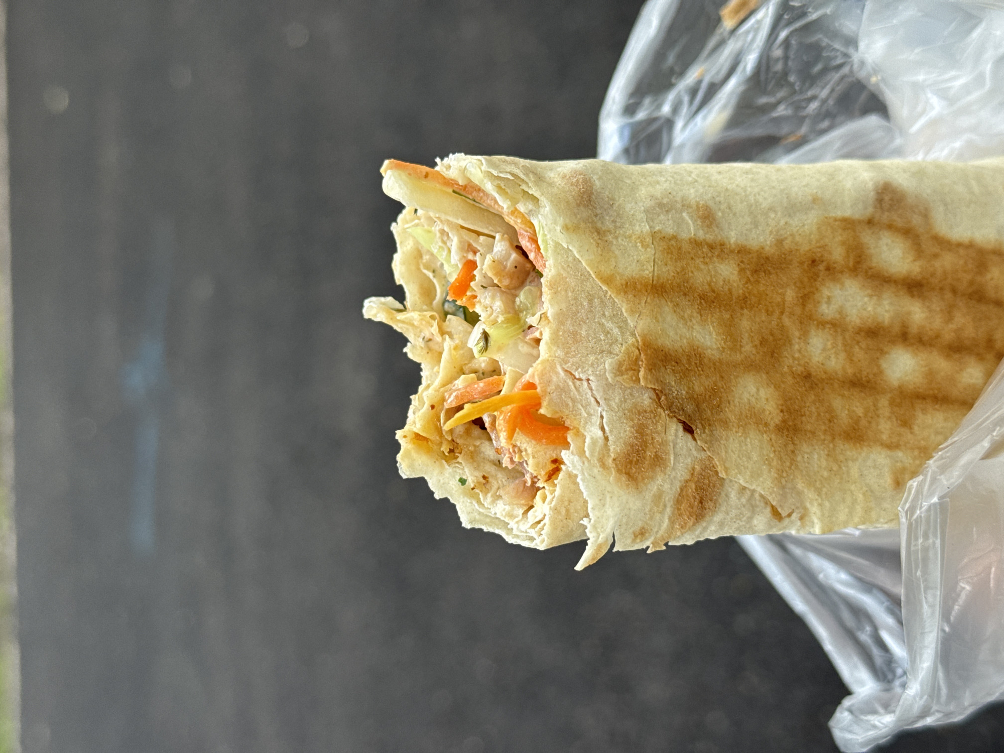 A close-up, vertical, eye-level shot shows a shawarma wrapped in a light brown, grilled tortilla. The top of the shawarma is slightly torn, revealing a filling of shredded chicken, thinly sliced carrots, and what appears to be a creamy sauce, along with some green herbs. The shawarma is held within a clear plastic bag. The background is a dark, textured surface, possibly asphalt, with a hint of light green at the very top. The lighting is natural, casting subtle shadows. The setting appears to be outdoors.