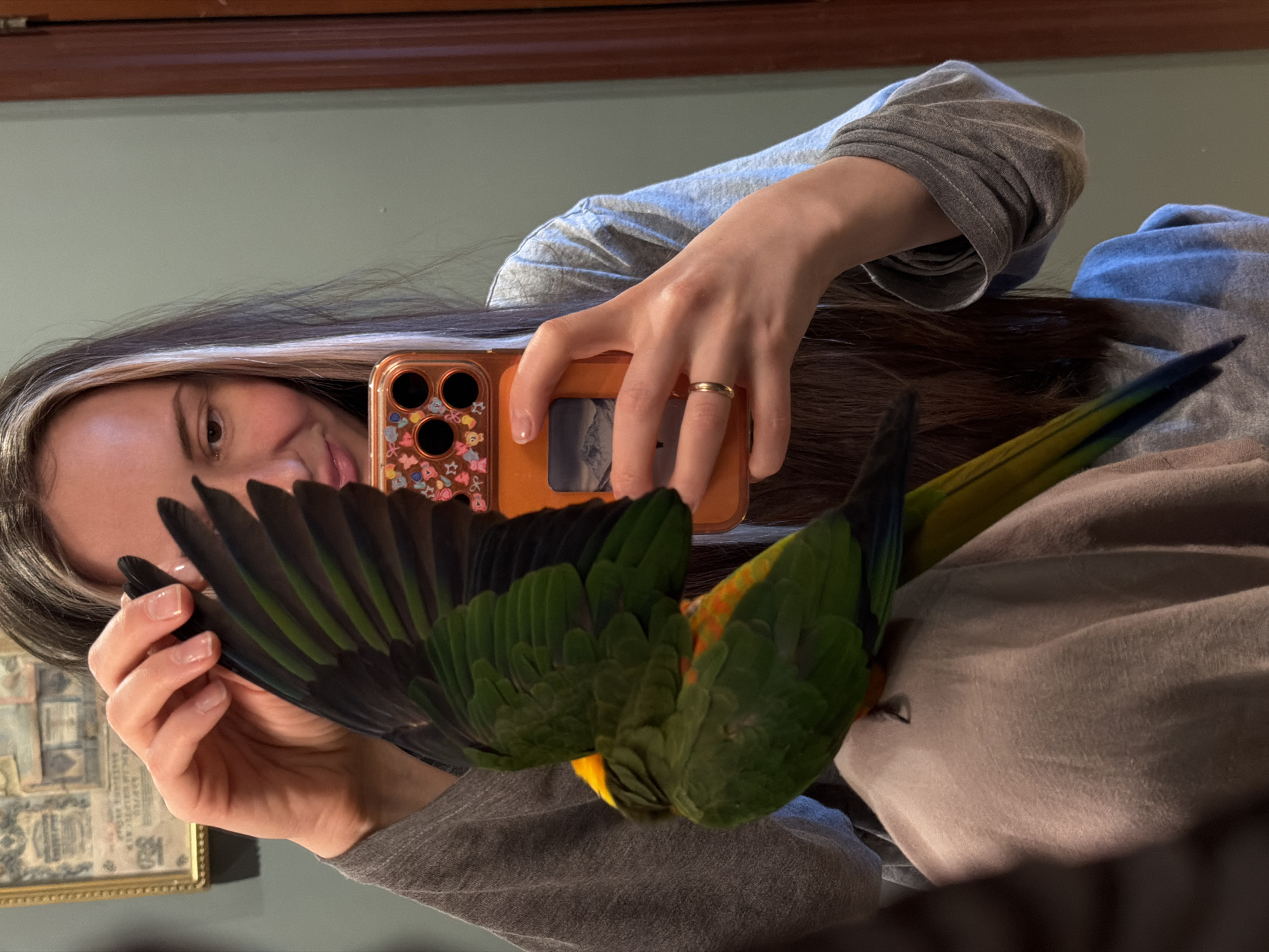 A woman holds a phone in her hands and takes a picture of a parrot. The woman has long dark hair with a streak of light hair. She wears a light blue shirt under a grey shirt, and a gold ring is visible on her finger. The phone has an orange case with a floral design. The parrot has green and yellow feathers. The location is not identifiable as Khimki, Russia from the image.