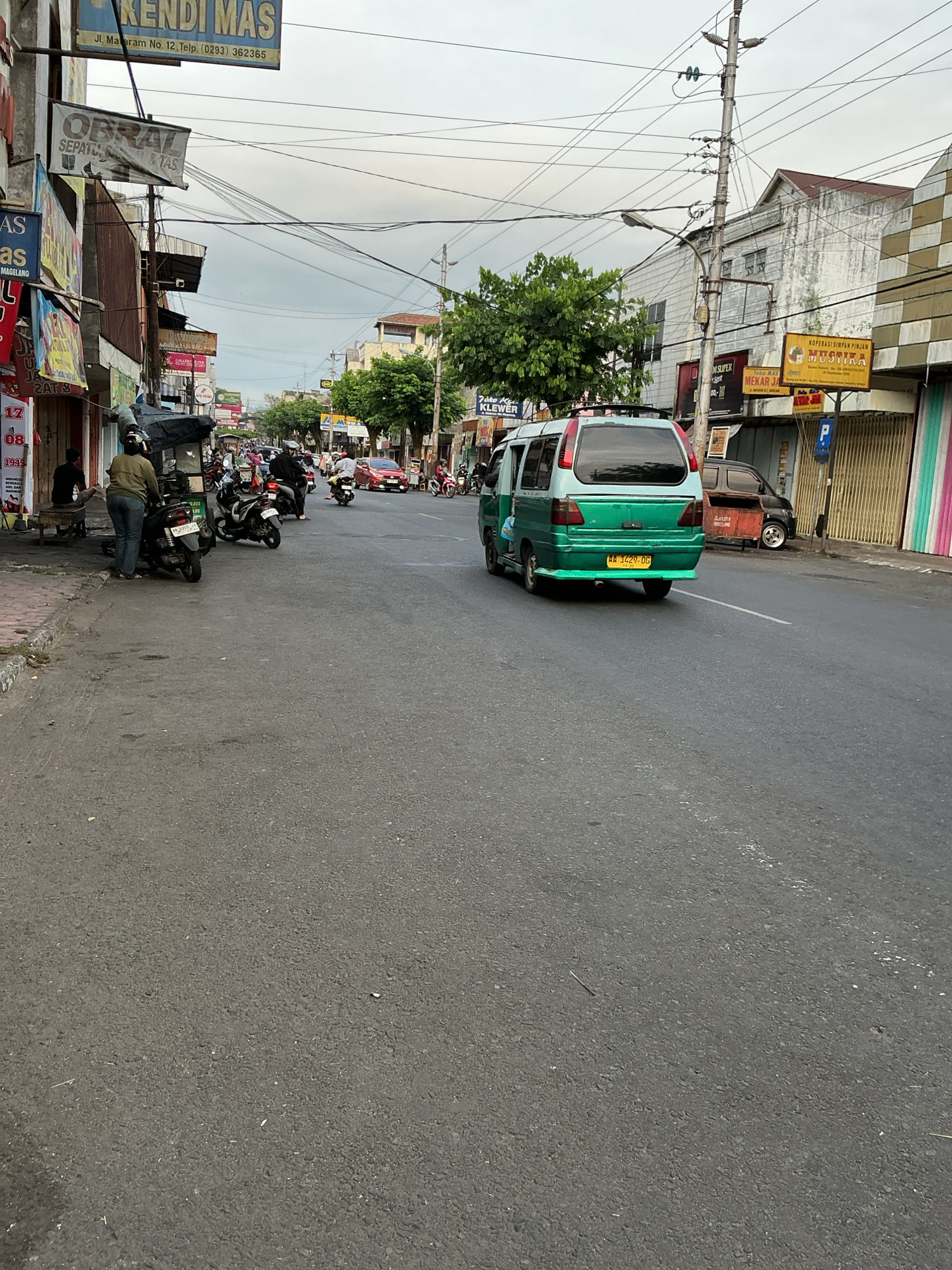 The image captures a street scene in Magelang, Indonesia, on an overcast day. The foreground is dominated by an asphalt road with a white line marking a lane. To the left, a sidewalk is visible with parked motorcycles and a few pedestrians. One individual, wearing a brown jacket and blue jeans, stands next to a parked motorcycle. Another person sits on a wooden bench near a storefront.
The middle ground features more parked motorcycles and a green and white van driving away from the camera. The van has a yellow license plate that reads "AA 1429 OG". Several other vehicles, including red cars and motorcycles, are visible on the road, suggesting moderate traffic.
The background reveals buildings lining both sides of the street. On the left, storefronts with various signs are present, including "KENDI MAS" and "OBRAL SEPATU & TAS". On the right, buildings display a more varied architectural style, with some featuring striped facades and others with older, weathered appearances. A prominent sign on the right reads "KOPERASI SIMPAN PINJAM MUSHIKA". Overhead, a complex network of electrical wires crisscrosses the sky. Trees are scattered along the street, providing some greenery. The overall atmosphere suggests a typical urban environment in Indonesia.