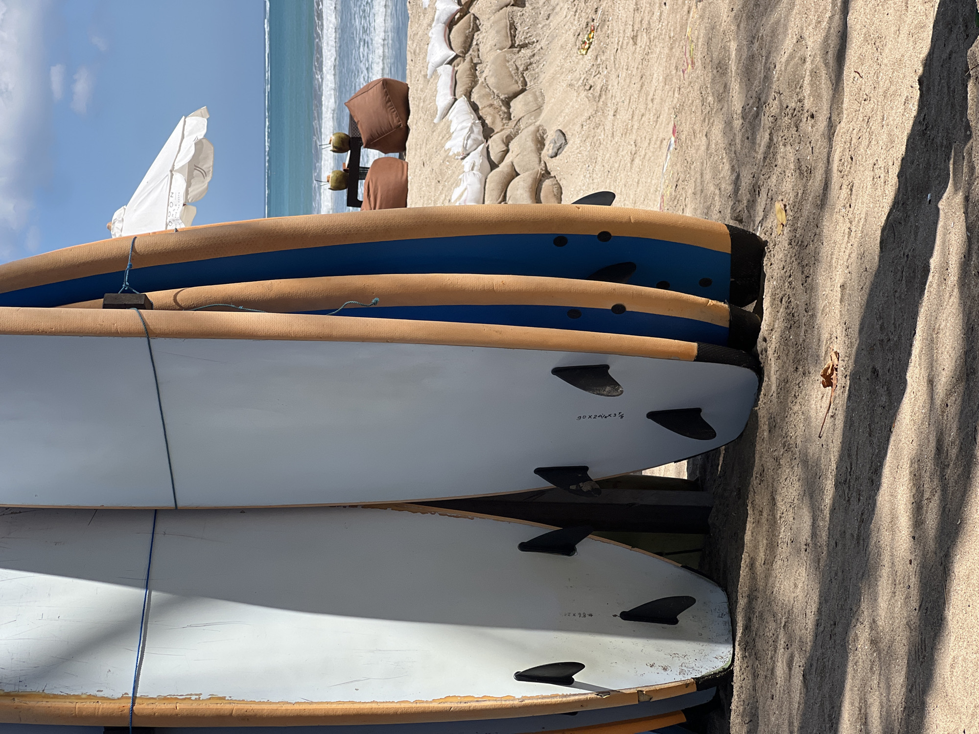 The image shows a row of surfboards standing upright on a sandy beach. The surfboards have white and orange foam exteriors, with blue accents on some. They are tied together with blue string. In the background, the turquoise ocean meets a clear blue sky. A white beach umbrella is open in the distance, and two coconuts with straws sit on a low table next to brown beanbag chairs. Sandbags form a barrier along the beach. The scene is likely Kuta, Bali, Indonesia, a well-known surfing destination.