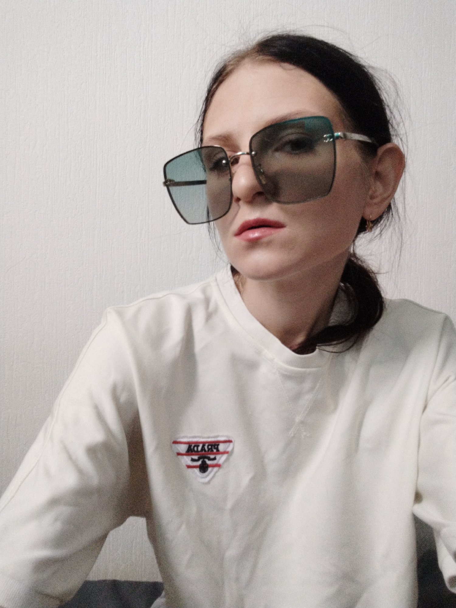 A woman wearing oversized square sunglasses, a white sweatshirt with a red, black, and white embroidered logo on the left chest, and a gold hoop earring on her right ear is pictured from the chest up, in an indoor setting with a textured white wall behind her. Her dark hair is pulled back into a ponytail. She is looking directly at the camera, with her mouth slightly open. The logo on her sweatshirt appears to be an inverted triangle with what looks like a mustache inside it, and the letters "ABYDY" are visible in red text at the top of the triangle. The lighting is soft and diffused, casting a slight shadow on the wall behind her. The overall image quality is somewhat grainy.