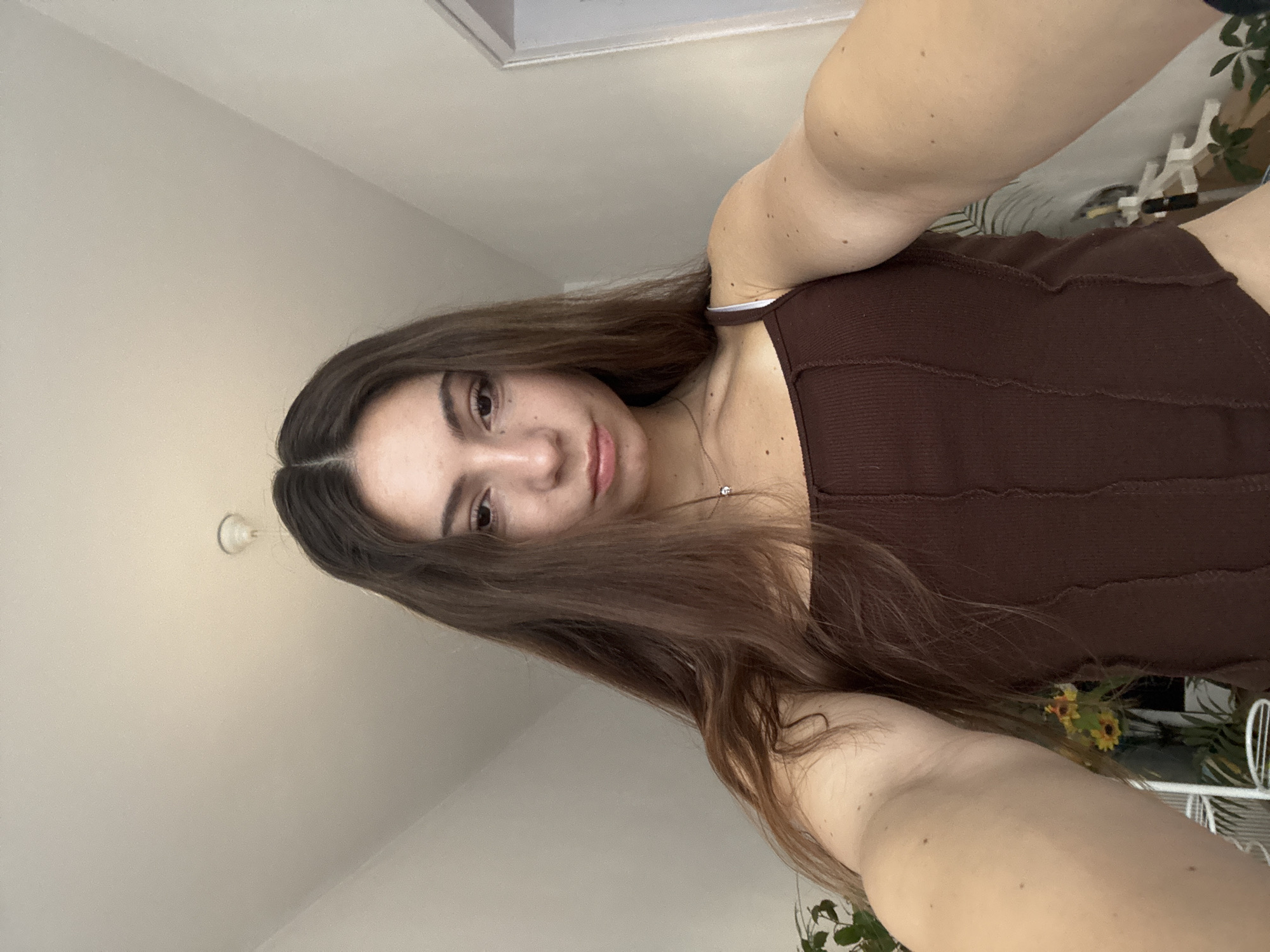 A young woman with long brown hair takes a selfie indoors. She is wearing a brown tank top. The background includes a ceiling, a picture frame, some plants and a white metal structure. It is not possible to determine the location of the city of Kaspichan, Bulgaria from this image.