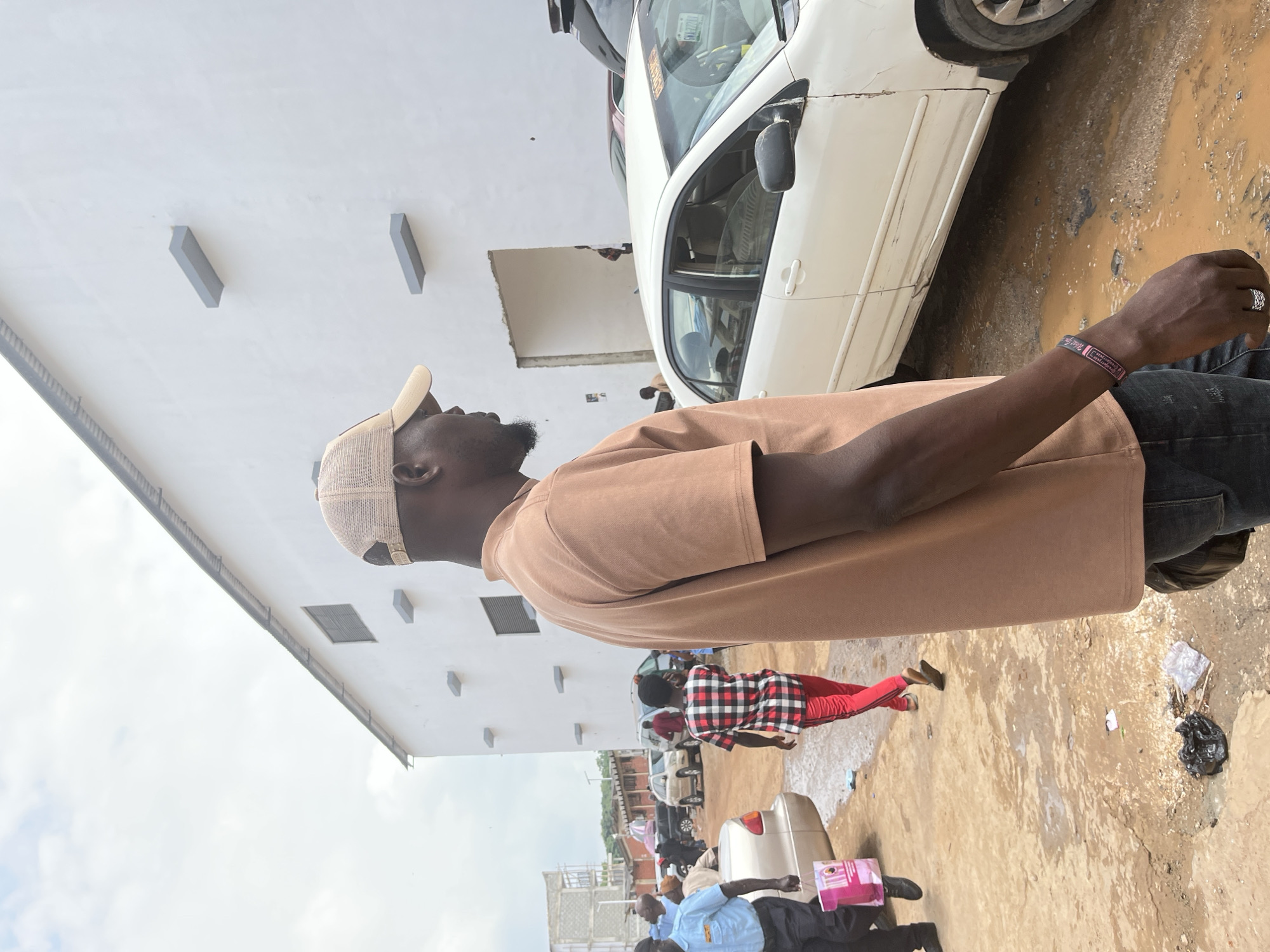 The image captures a street scene in Abuja, Nigeria. In the foreground, a man wearing a tan baseball cap and a tan t-shirt is positioned in profile, facing to the left. He has a beard and is wearing dark jeans and a ring on his left hand. A bracelet is visible on his right wrist. Behind him, to the left, is a white building with a dark-lined corrugated roof and several rectangular gray protrusions on its white exterior wall. The ground is unpaved, with visible puddles of water and dirt. Several cars are present in the background, including a white sedan with visible damage to its side, a silver sedan, and other vehicles obscured by distance and other subjects. People are scattered throughout the scene, some walking and others standing near vehicles. One man in the middle ground is wearing a red and black checkered shirt and red pants, walking away from the camera. Another person, possibly a security guard in a light blue uniform, is walking towards the left foreground carrying a pink shopping bag. The sky is overcast.