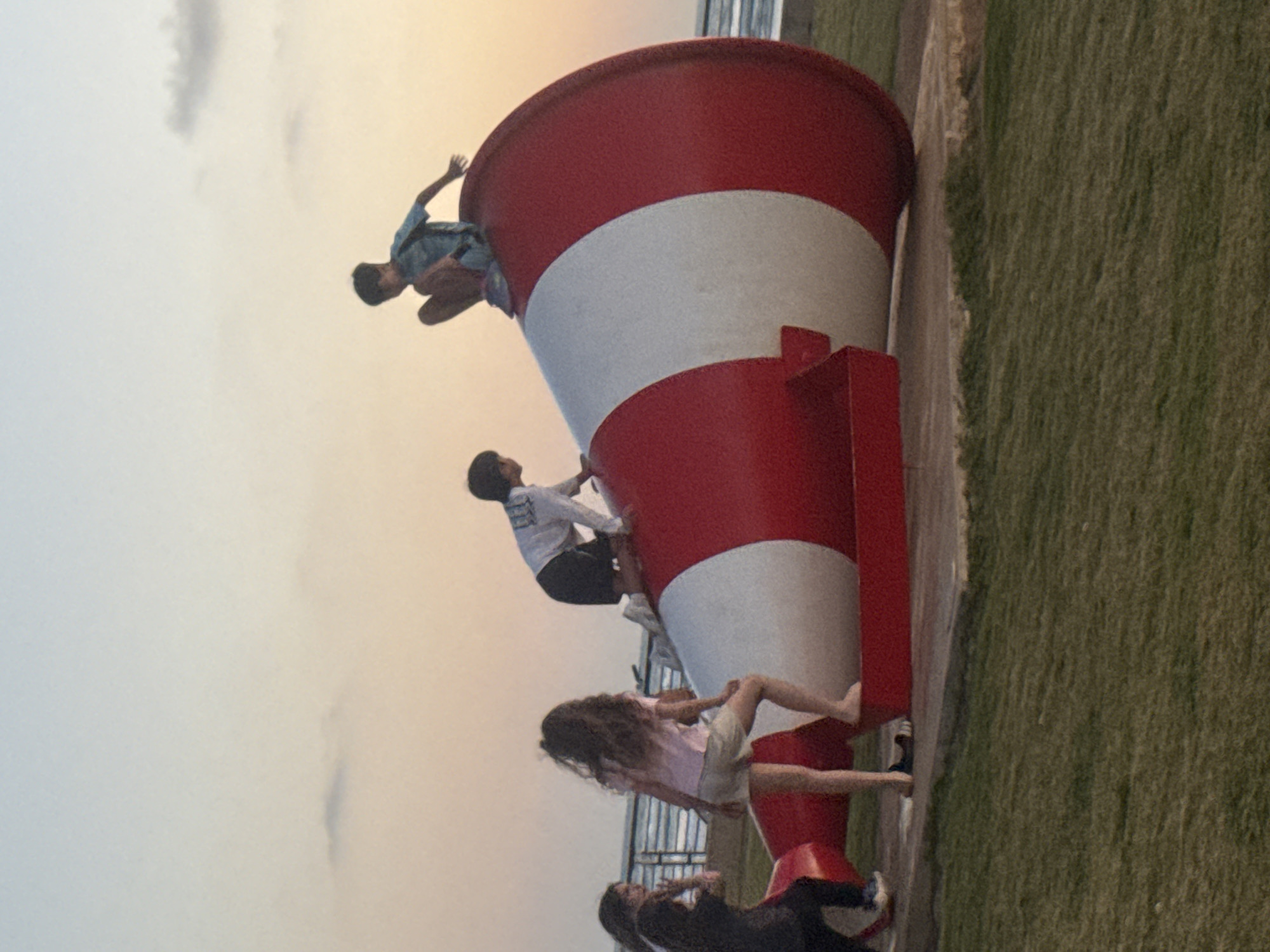 A group of four people are climbing on a large, red and white striped megaphone-shaped structure. The structure sits on a paved surface next to a grassy area. The sky is overcast. This megaphone structure is located near the beach in Tel Aviv, Israel.