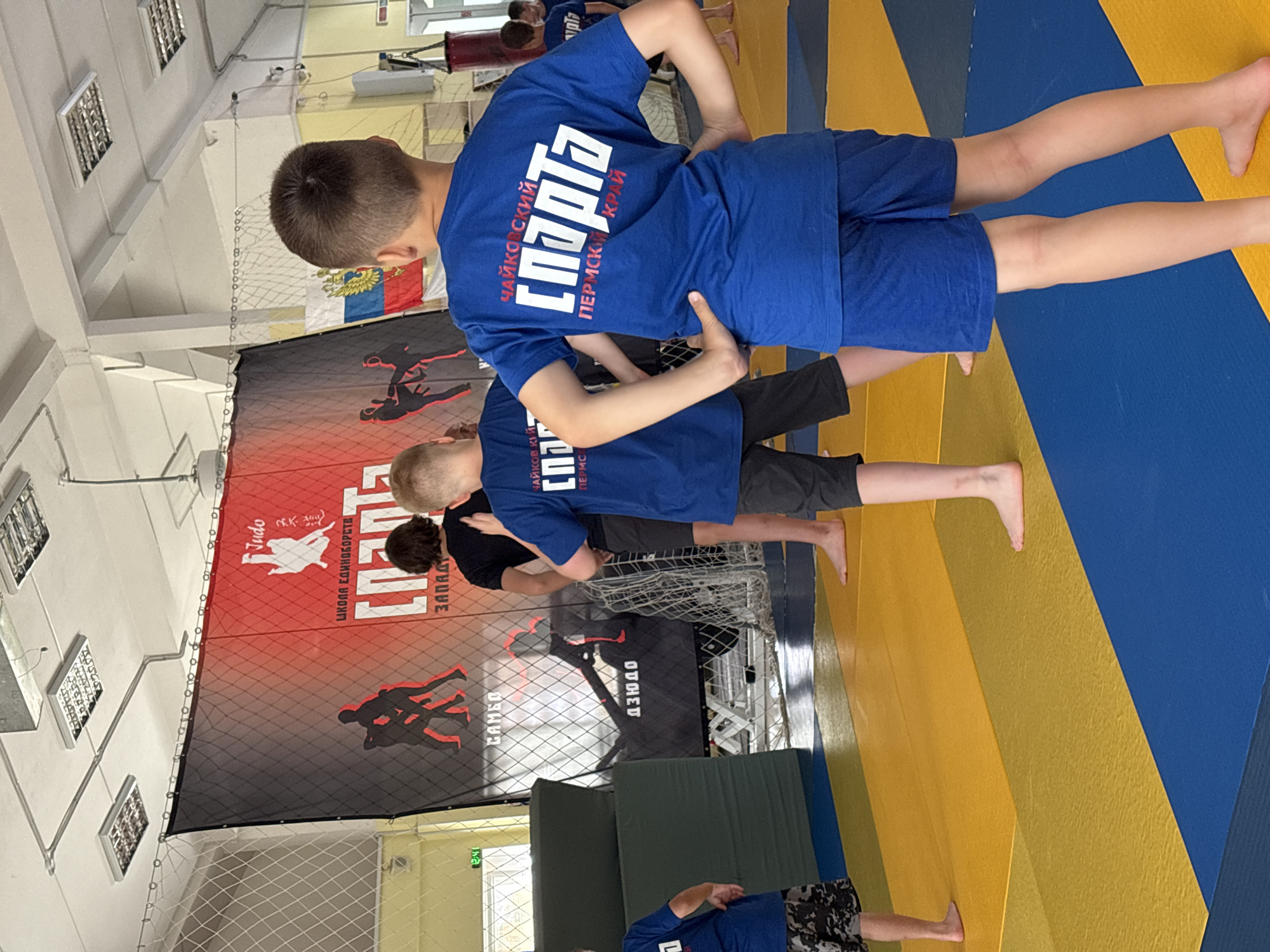 The image depicts an indoor training facility with a judo mat on the floor. Several children, dressed in athletic wear, are present on the mat, seemingly participating in a judo or martial arts class. In the background, a large banner with red and black colors displays the word "Judo" and some silhouettes of judokas. The banner also features text in Russian, including "ШКОЛА ЕДИНОБОРСТВ" (School of Martial Arts) and "СПАРТА" (Sparta). A Russian flag is visible behind the banner. The environment suggests a sports school or club. The image appears to have been taken in the city of Chaikovsky, Russia, as indicated by the text on the children's shirts, which reads "Чайковский" (Chaikovsky) and "Пермский край" (Perm Krai).