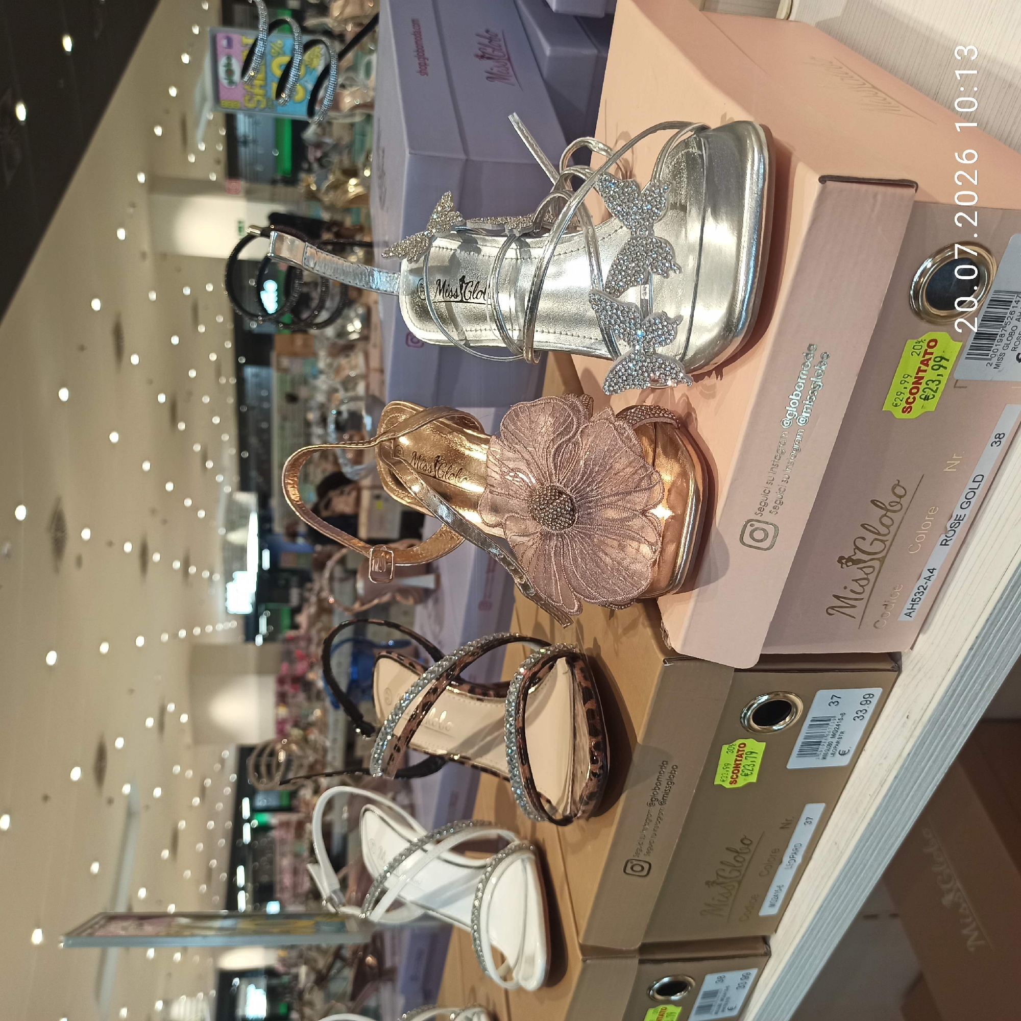 This is a medium shot, eye-level, indoor photograph of a retail display of women's high-heeled sandals in what appears to be a shoe store or department store. The shoes are arranged on top of their original packaging boxes. The background shows more shelves stocked with shoes and boxes, as well as ceiling lights and some blurry figures of shoppers.
The foreground features four pairs of open-toed high-heeled sandals. From left to right:
1. A white sandal with thin, criss-crossing straps adorned with rhinestones and a rhinestone-covered heel strap.
2. A sandal with a leopard print strap across the toes and a leopard print and tan criss-crossing ankle strap, also embellished with rhinestones.
3. A rose gold sandal with a large, layered, sequined flower embellishment on the toe strap.
4. A silver sandal with multiple thin straps, decorated with three sparkly butterfly embellishments.
The shoes are displayed on cardboard boxes. The boxes have branding and product information printed on them. The box for the white sandal is partially visible on the left, with a white price tag. The box for the leopard print sandal is to its right, with "Miss Globo" branding and a yellow sticker indicating a sale price of €23.79 from €33.99. The box for the rose gold sandal is pink with "Miss Globo" branding and a similar yellow sale sticker indicating €23.99 from €29.99. The box for the silver sandal is also pink and visible on the far right, with some text and a date stamp in the bottom right corner.
The lighting is bright and even, suggesting good store illumination. The overall impression is one of a well-organized retail environment showcasing fashionable footwear. The image quality is clear, allowing for detailed observation of the shoes and their packaging. The timestamp in the bottom right corner reads "20.07.2026 10:13". The store is identified as Miss Globo, a footwear retailer, with social media handles suggested on the boxes, including Instagram accounts @globomoda and @missglobo. Based on the social media information, the store is likely located in Bari, Italy.