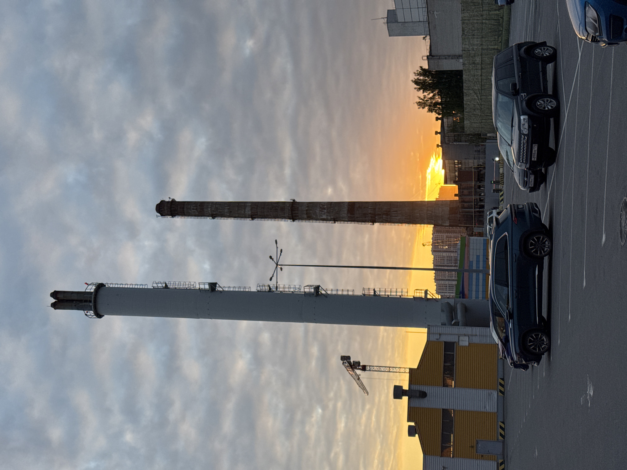 The image captures an industrial area at sunset, with a prominent orange glow from the setting sun on the right side of the frame. The sky transitions from a soft grey-blue on the left to vibrant orange and yellow hues on the right, interspersed with scattered clouds.
Two tall industrial chimneys dominate the scene. On the left, an older, reddish-brown chimney shows signs of age. To its right, a newer, light grey chimney stands, featuring visible ladders and platforms wrapping around its structure, with multiple vents at its top.
In the foreground, a paved parking lot occupies the lower half of the image. Several cars are parked, including a dark-colored SUV, likely a Land Rover, prominently displaying the license plate "H898HP 178". This region code "178" corresponds to Saint Petersburg, Russia. Another dark SUV is partially visible closer to the viewer, and a blue car is seen in the upper right corner of the parking lot.
Behind the chimneys and cars, various industrial buildings are present, some illuminated by the warm light of the setting sun. One building features distinctive yellow and black stripes. A construction crane is visible in the mid-ground. In the far distance, the silhouette of a city skyline can be seen against the bright horizon. A multi-lamp street light pole is also visible between the chimneys. The scene depicts a quiet moment in an industrial or utility complex on the outskirts of Saint Petersburg, Russia, as the day ends.