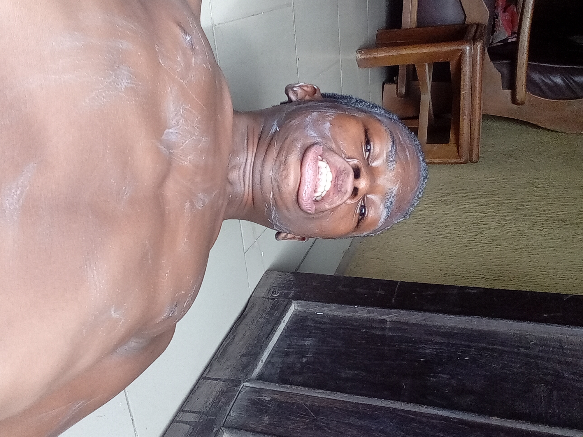 A man is looking at the camera and smiling, the man has white cream smeared on his face and torso. He is in what appears to be a room in a house. There is a tiled floor and a wooden table in the foreground. In the background are wooden chairs. This scene is likely taking place in Onitsha, Nigeria, but that information cannot be confirmed with certainty from the image alone.
