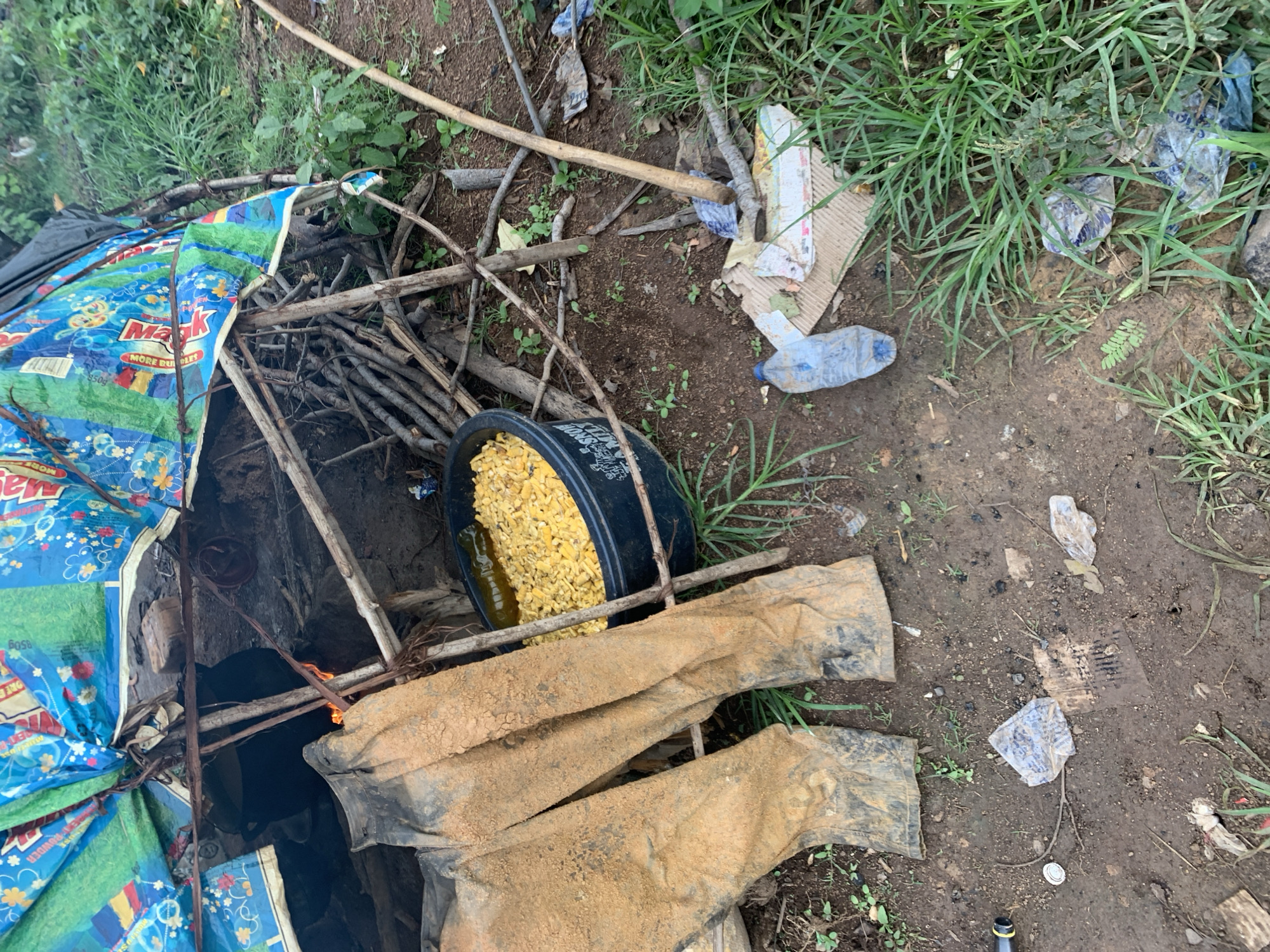The image depicts an outdoor scene with elements suggestive of a temporary dwelling. In the foreground, a pair of dirty, mud-caked trousers are laid out on the ground. Behind the trousers, a large black basin filled with yellow kernels, possibly corn, is visible. The dwelling itself appears to be constructed from branches and covered with a colorful, patterned tarp. Debris, including plastic bottles and cardboard, is scattered around the immediate area, which consists of dirt ground and overgrown grass. The overall impression is one of a makeshift campsite or informal settlement.