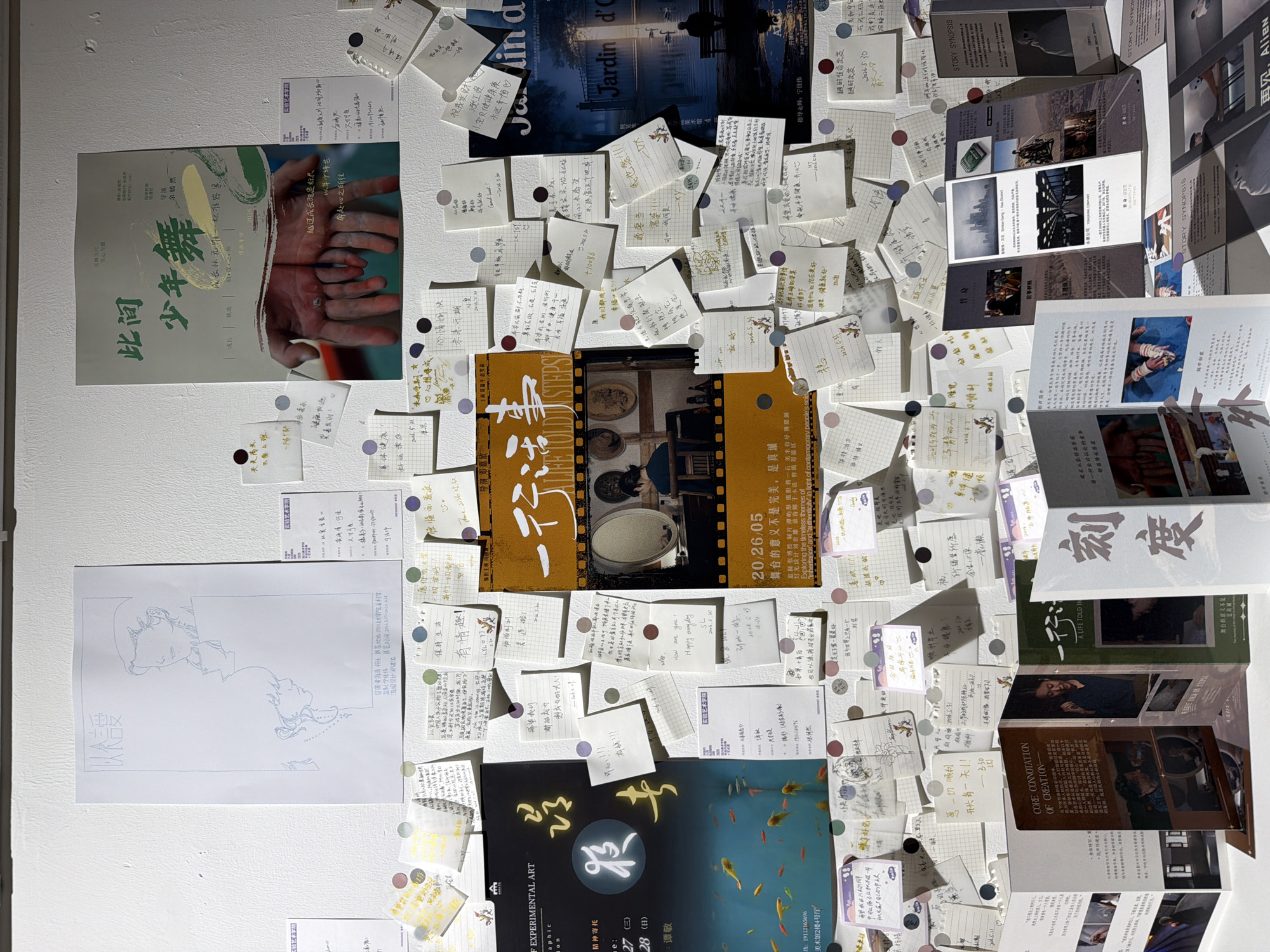 This image captures a wall display, likely part of an exhibition or bulletin board, situated in the Chongqing High-tech Zone, China. The wall is predominantly white, covered with various posters, numerous handwritten sticky notes, and several folded brochures arranged along the bottom edge.
At the top left, a light blue line drawing poster depicts two stylized figures, one above the other, with Chinese characters "以愛" (With Love) at the top. To its right, another poster titled "此間少年舞" (This Young Dance) features an image of two intertwined hands with calluses, set against a green and white background.
In the mid-section on the left, a dark blue poster shows swimming goldfish with the character "歸" (Return/Belong) in a yellow circle. Adjacent to it, a central poster titled "一行故事" (A Life Told in Steps) displays an oval mirror reflecting a woman from behind, with text including "20 26 05" and "Exploring the timeless themes of inheritance and authenticity." To the far right, a dark blue poster with "Jardin de" visible at the top shows a solitary figure on a bench beside a lake.
The central and lower portions of the wall are densely covered with dozens of small, square sticky notes, primarily white, light yellow, or grid-patterned. Most notes feature handwritten messages in Chinese, some with dates like "2024.5.30" or "June 2024." These notes are affixed with small, round, colorful translucent stickers. The messages appear to be comments, feedback, or wishes, with snippets like "How are you? Happy everyday" in English on one note.
Along the bottom of the display, several multi-fold brochures are fanned out. One prominently displays the title "刻度" (Scale/Degree) in large characters. Other brochures feature images of individuals, cityscapes, or abstract designs, with some English titles such as "CORE CONNOTATION OF CREATION," "A LIFE TOLD IN STEPS," and "STORY SYNOPSIS." The overall scene suggests an interactive public display, possibly for an art or film exhibition, encouraging visitor engagement.