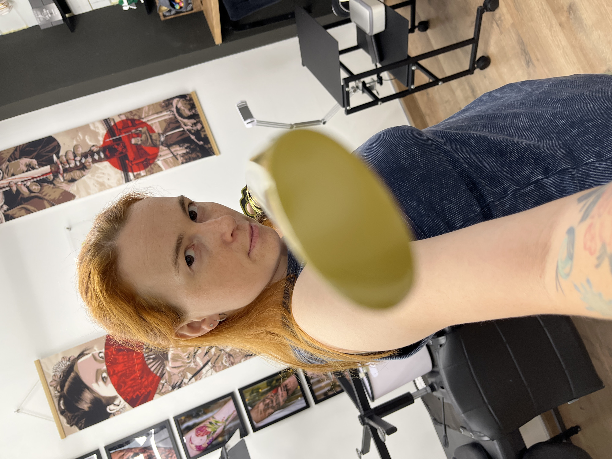 A Caucasian woman with shoulder-length auburn hair and fair skin is in a brightly lit room that appears to be a tattoo parlor, indicated by the tattoo equipment and artwork on the walls. She is holding a round, yellow object out towards the camera, which is blurred and out of focus, obscuring part of her face. She is wearing a dark blue, ribbed top. Behind her, a large, vertical tapestry with an image of a person holding a sword and a red circle is hanging on the wall. To her left, several framed pictures of tattoos are displayed. To her right, a black rolling stand with a table is visible, along with a tattoo machine and other supplies. The floor is made of light-colored wood. The image is a close-up selfie taken from a slightly low angle. The location in the city of Chaikovsky, Russia, is not discernible from the image.