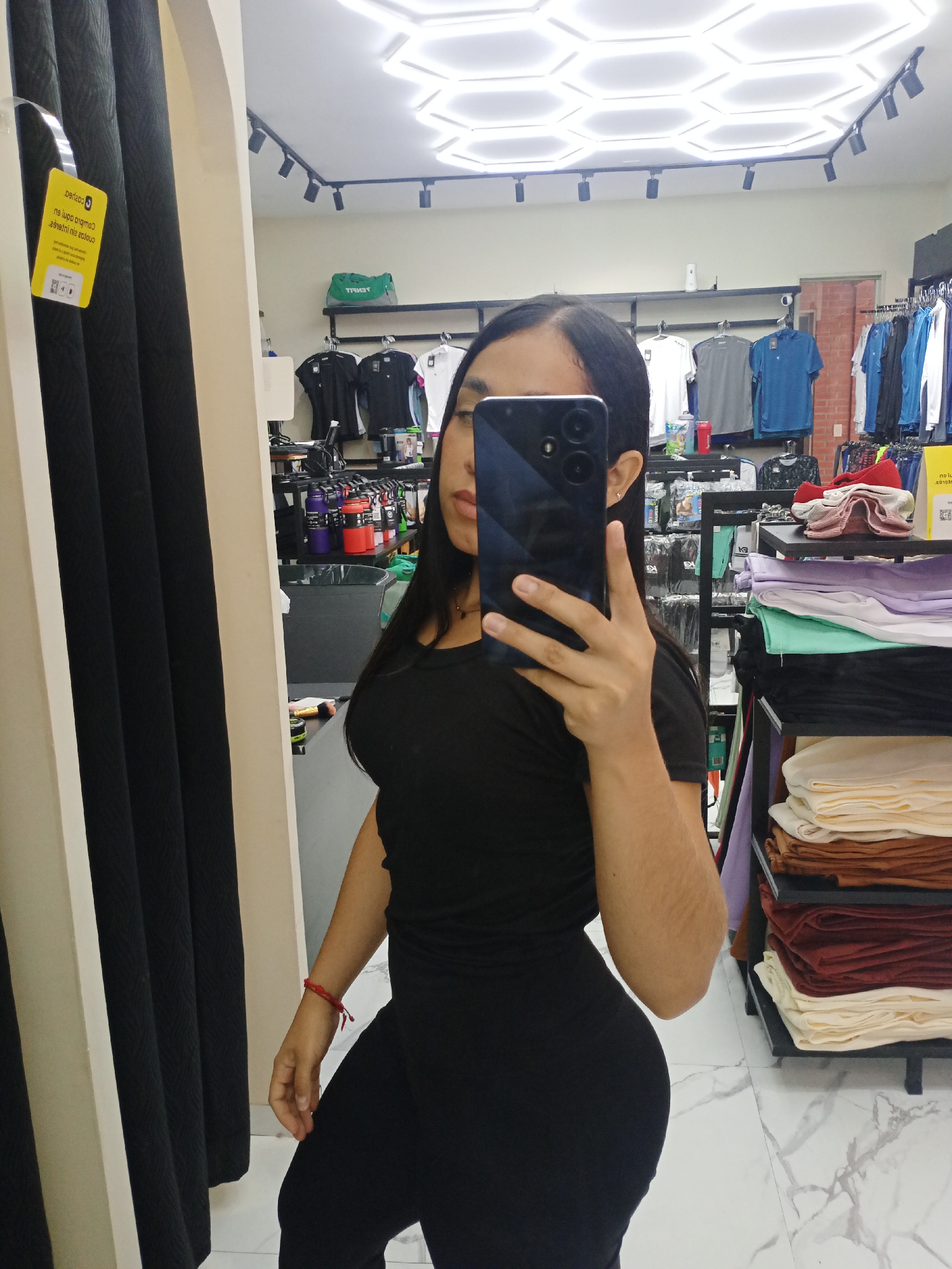 A person is taking a mirror selfie inside what appears to be a retail clothing store. The individual is wearing a form-fitting black top and black leggings, with long dark hair partially visible around the dark-colored smartphone used to capture the image. A red string bracelet is worn on the right wrist.
The background, seen through the mirror's reflection, reveals various elements of the store. Clothing racks hold a selection of shirts in black, white, gray, and blue. To the right, shelves display neatly folded textiles or apparel in colors such as white, cream, light green, purple, black, brown, and red. In the middle-left, a shelf contains several colorful water bottles or containers, and a black trash can is also visible. Above these, a green shopping bag with the mirrored word "VENATT" is placed on a shelf.
The store's ceiling features modern hexagonal light panels and black track lighting with spotlights. The floor is covered with white and grey marble-patterned tiles. On the left side of the mirror, dark, textured curtains or fabric samples hang, adjacent to the mirror's light-colored frame. A yellow tag is attached to these curtains, displaying mirrored text, including "perlaso," phrases like "ne luge omgua" and "s916ilni zolouɔ," and a QR code symbol. The scene depicts a well-lit, organized retail environment.
