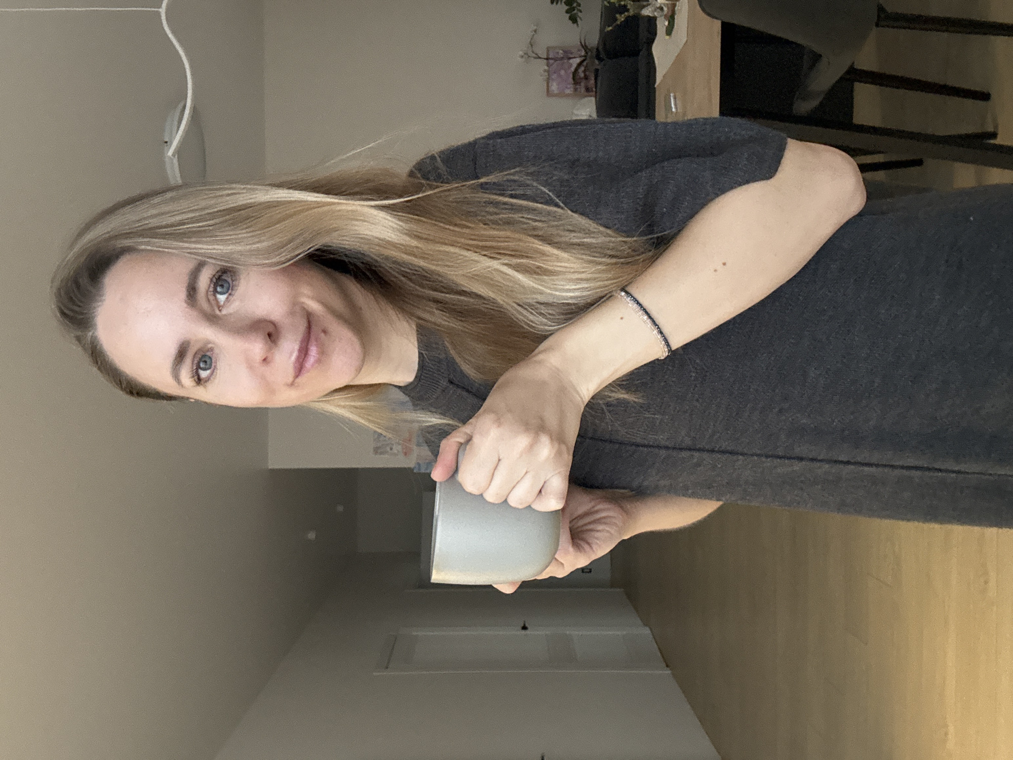 An image captured indoors features a woman with long, wavy blonde hair and blue eyes. She is looking directly at the viewer with a gentle, soft smile. She wears a dark grey, long-sleeved top or dress and a delicate beaded bracelet on her left wrist. Both hands are clasped around a light grey, ceramic mug, held at her mid-chest level.
The background depicts a modern home interior, with light beige or off-white walls and a warm wooden floor visible in the lower right. Blurred elements in the distance suggest household furniture, including a dining table with dark chairs and what might be shelves or a cabinet adorned with greenery. A white cable or light fixture hangs from the ceiling in the upper left corner. The lighting is soft and diffused, creating a relaxed atmosphere, consistent with an indoor daytime setting. The woman's expression conveys a sense of calm and friendliness. No visible text is present in the image. The location context provided is Chaikovsky, Russia.