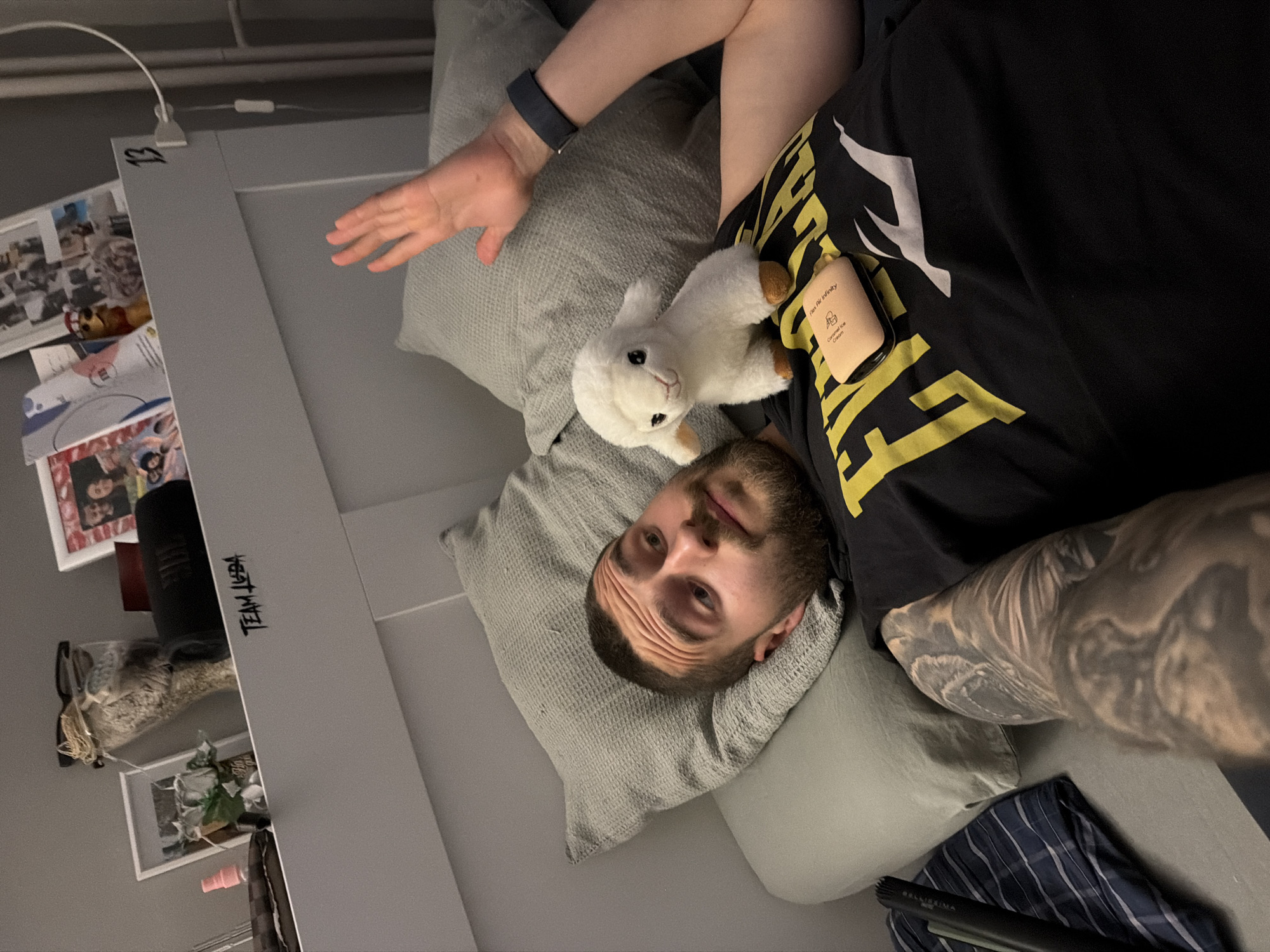 An overhead shot, rotated ninety degrees clockwise, shows a man lying on what appears to be a bed or sofa in an indoor setting in Sofia, Bulgaria. The man, with a short beard and dark hair, looks directly at the camera with a neutral expression. He is wearing a black t-shirt with yellow and white graphics and has extensive tattoos on his right arm. A black smart watch is visible on his left wrist.
Resting on his chest is a small white stuffed lamb toy with black eyes. Next to the lamb, a small, light brown rectangular container is visible, bearing the text "Flex Air Infinity" and "Coconut Ice Cream" with a small ice cream cone icon. The man's head rests on a textured grey pillow, with another similar pillow partially visible under his arm.
To his left (viewer's right, if rotated to be upright), a dark grey headboard or panel features handwritten text: "13" and further down, "TEAM JUDA". A white bendable lamp is clipped to the top of this panel. Below the lamp, on a shelf, several framed photographs are displayed along with a dark cylindrical object (possibly a speaker), a small pink bottle, and various other personal items. Part of a blue and white striped blanket is visible in the bottom right corner of the frame. A black marker pen, with "SALLIEKIMA" printed on it, lies near the blanket. The lighting in the room appears to be artificial.