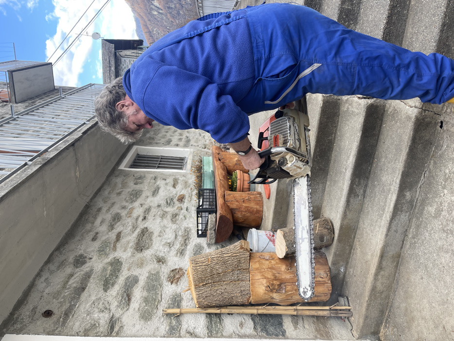 An older man with gray hair, dressed in a blue fleece top and blue overalls, is captured mid-action, focused on cutting a large log of wood with a chainsaw. He is bent forward over the log, which rests on one of the concrete steps of a stairway. The chainsaw, clearly branded "Stihl," has a long metal bar and chain, with wood chips visible from the ongoing cut.

The setting is outdoors, on a set of concrete stairs leading upwards. To the man's left, a traditional stone wall features a small rectangular window secured with a metal grill. Below the window, a rustic, thick wooden bench or table is visible, holding a potted plant. Several other logs, some stacked, along with a white bucket and black plastic crates, are arranged around the cutting area. Bamboo poles lie horizontally at the base of the wood pile.

In the background, under a bright blue sky dotted with scattered white clouds, other stone buildings with tiled roofs can be seen, leading up to distant mountains. The clear daylight suggests it's a sunny day. The scene depicts a practical chore in a picturesque, mountainous village environment, specifically located in Dosso (Ciappanico), Italy.