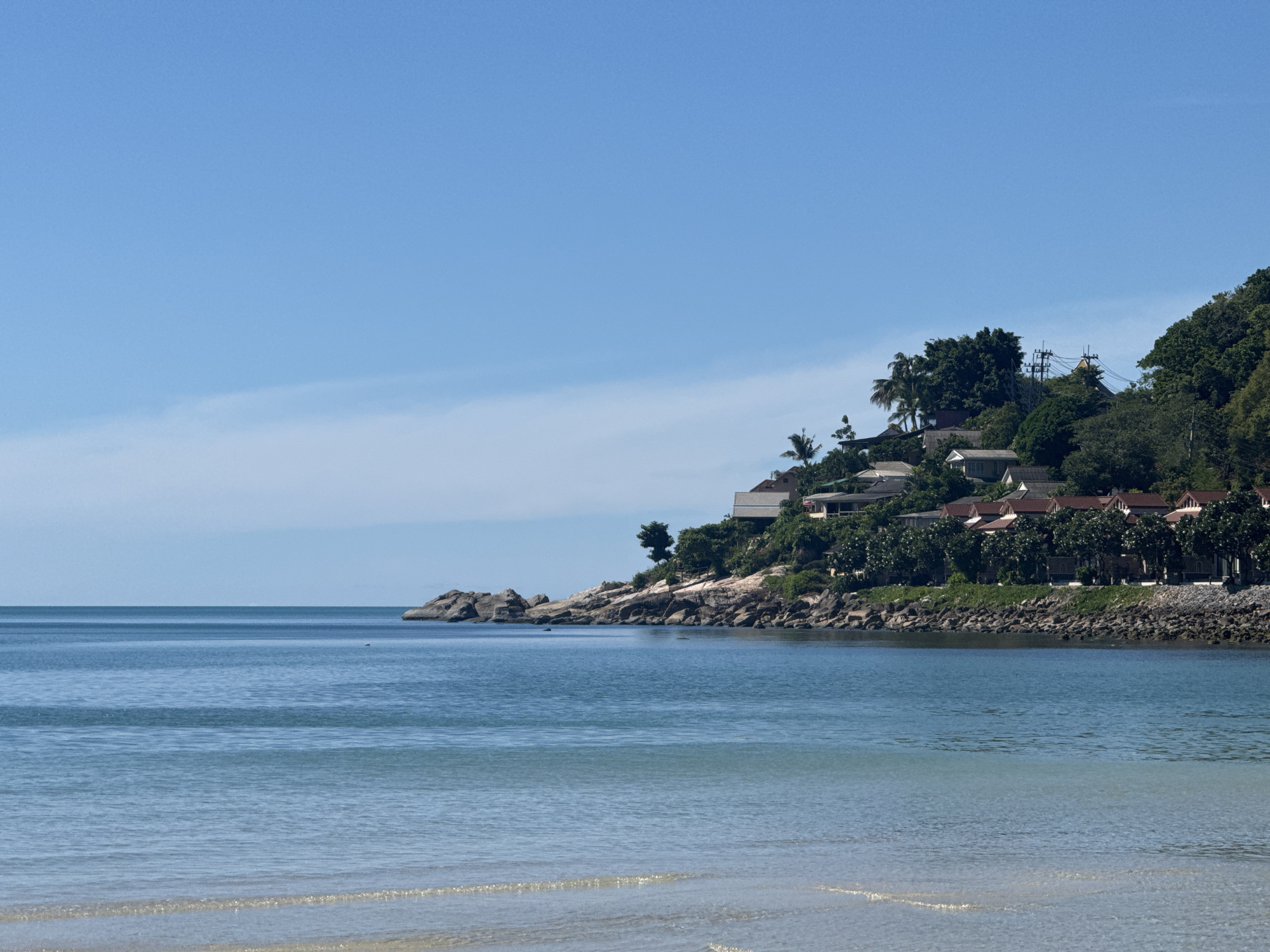 The image depicts a coastal scene in Baan Chaweng Noi, Thailand. The foreground consists of clear, shallow ocean water with gentle ripples reflecting the bright sky. In the midground, a rocky shoreline juts out into the sea, with several large boulders scattered along the coast. Behind the rocks, a lush green hillside rises, densely covered with trees and palm trees. Several buildings with red-tiled roofs are nestled into the hillside, appearing to be part of a resort or residential area. Some buildings are visible closer to the water, while others are higher up on the slope. Utility poles and wires are also visible amongst the trees. The sky above is a clear, vibrant blue with a few wispy white clouds. The overall impression is a serene tropical landscape under a sunny day.