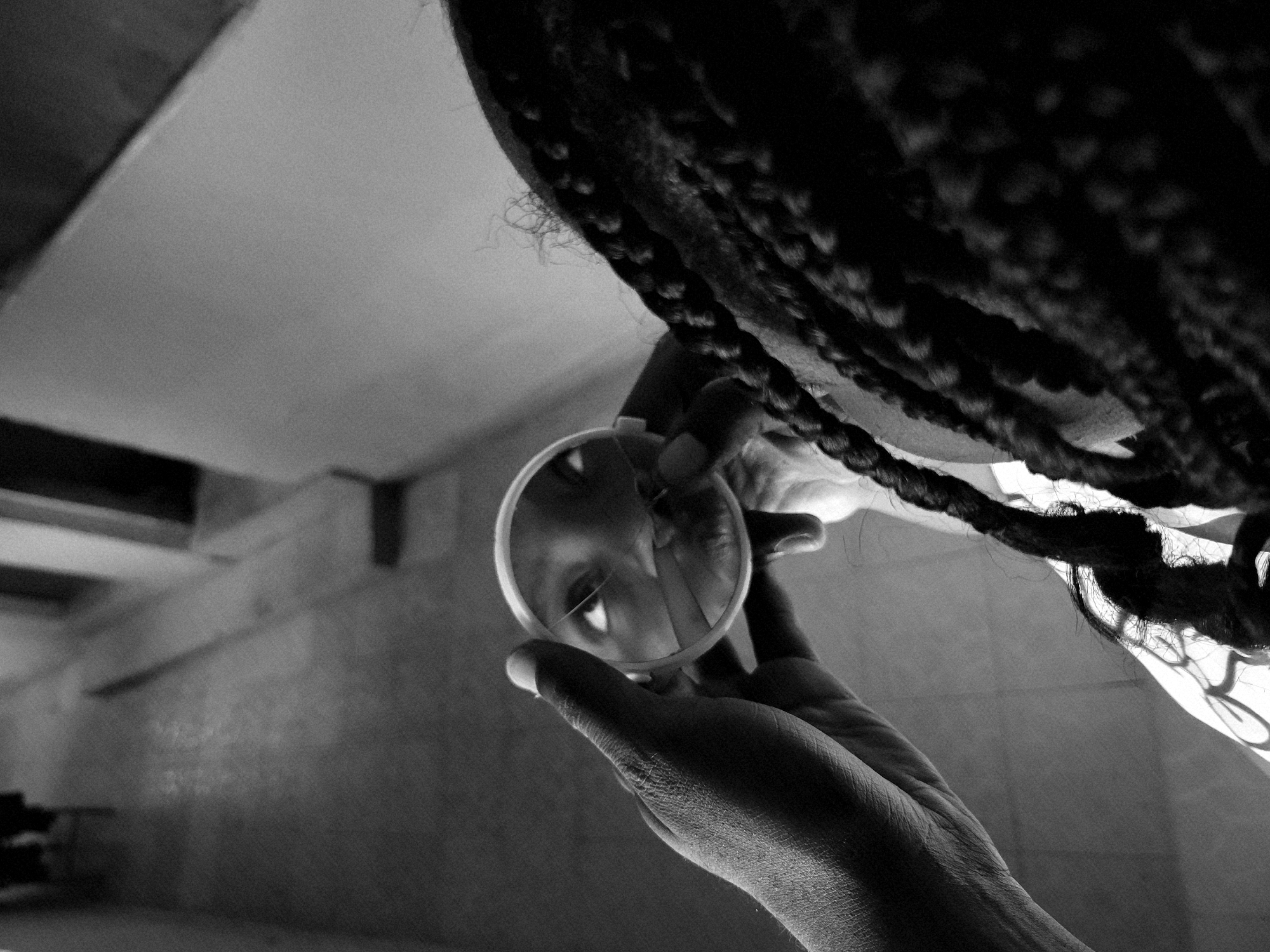 A black and white, high-angle photograph shows a person holding a small, round mirror reflecting their eye. The mirror appears to have a crack in it. The person has dark hair styled in braids that drape over the upper right portion of the image. The left side of the photograph features a blurred interior scene, possibly a hallway with furniture and architectural details, including a ceiling and wall. Without additional information, pinpointing the location to Jos, Nigeria, is not possible based solely on the visual details.