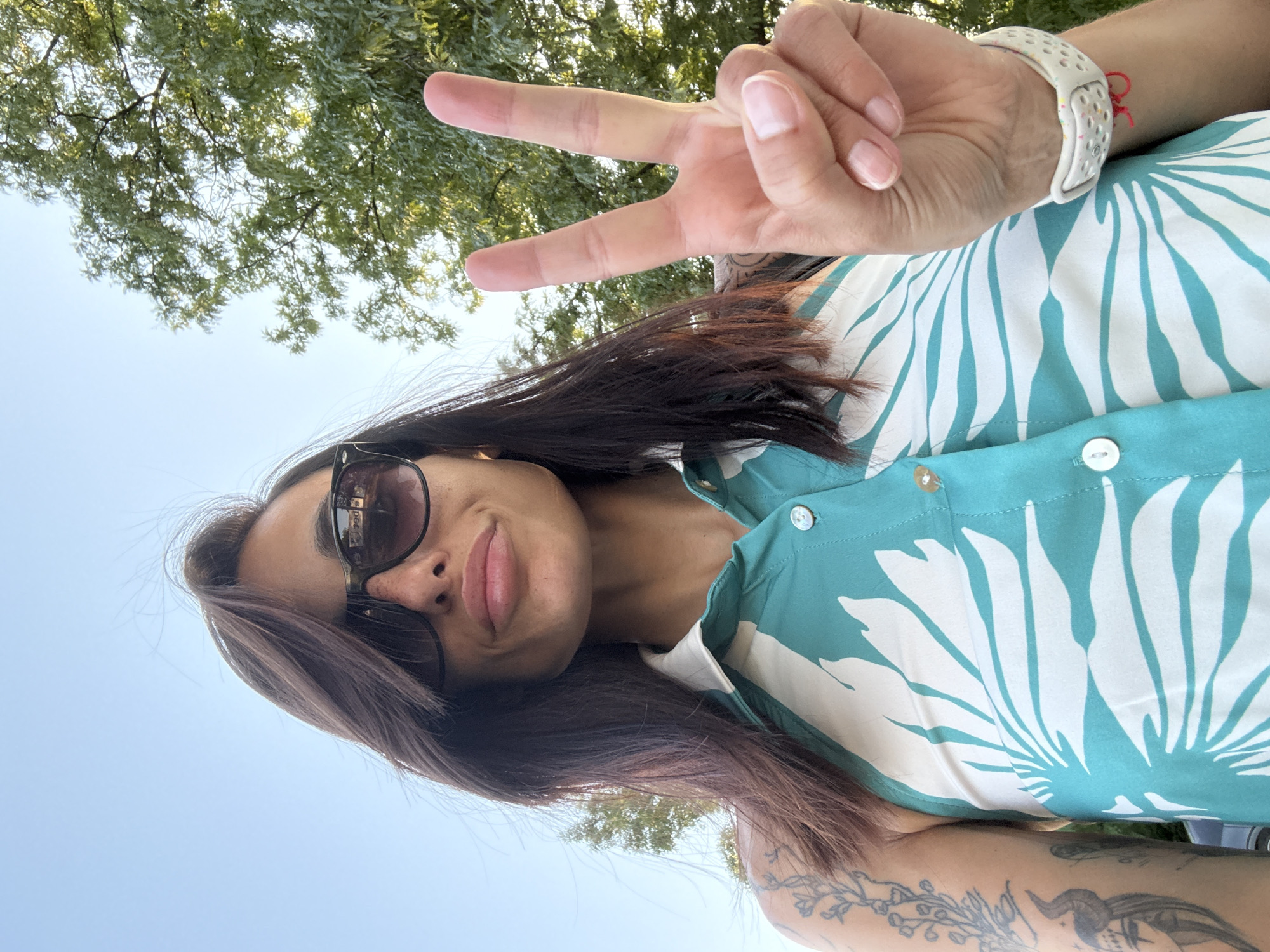 The media file is a selfie taken outdoors, likely during the daytime, given the bright sky and natural lighting. The subject is a woman with dark hair, wearing sunglasses and a teal and white floral print shirt. She is making a peace sign with her right hand, which also shows a white wristwatch with a speckled band and a red string bracelet. The background features green foliage from trees and a clear sky. There are tattoos visible on her arm. The image is oriented vertically, with the top of her head towards the bottom right of the frame. The overall mood appears casual and cheerful, suggesting a relaxed outdoor activity. There is no visible text in the image.