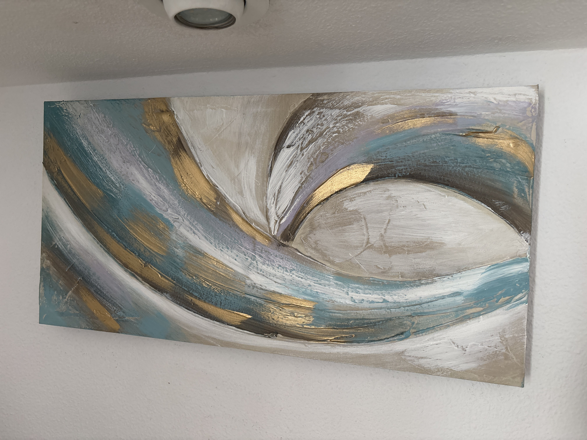 A horizontal abstract painting hangs on a white textured wall beneath a white ceiling with a circular light fixture. The painting features thick, textured brushstrokes in shades of white, beige, light purple, teal, and gold, creating a dynamic, swirling pattern. The gold elements appear to be metallic paint, catching the light. The painting is centered on the wall and extends across most of its width. The environment appears to be indoors, likely a residential or office space. No people are visible in the image. The location in Orihuela, Spain, is not discernible from the image itself.
