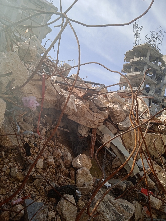 The image depicts an urban landscape in Khan Yunis, Palestinian Territory, characterized by extensive structural damage under a bright, partly cloudy sky. The foreground and midground are filled with a large volume of rubble, primarily composed of irregularly shaped concrete blocks, broken masonry, and numerous twisted, rusted rebar rods. These rebar elements are bent into various configurations, some forming prominent curves. A crumpled pink plastic bag is visible among the debris on the left side of the frame. In the background, to the right, stands a multi-story concrete building exhibiting severe structural failure, with multiple floors partially collapsed, walls missing, and internal frameworks exposed. The top of this damaged building features what appears to be communication tower structures, including antennas and scaffolding. No people or active interactions are present in the scene. The overall environment indicates widespread destruction.