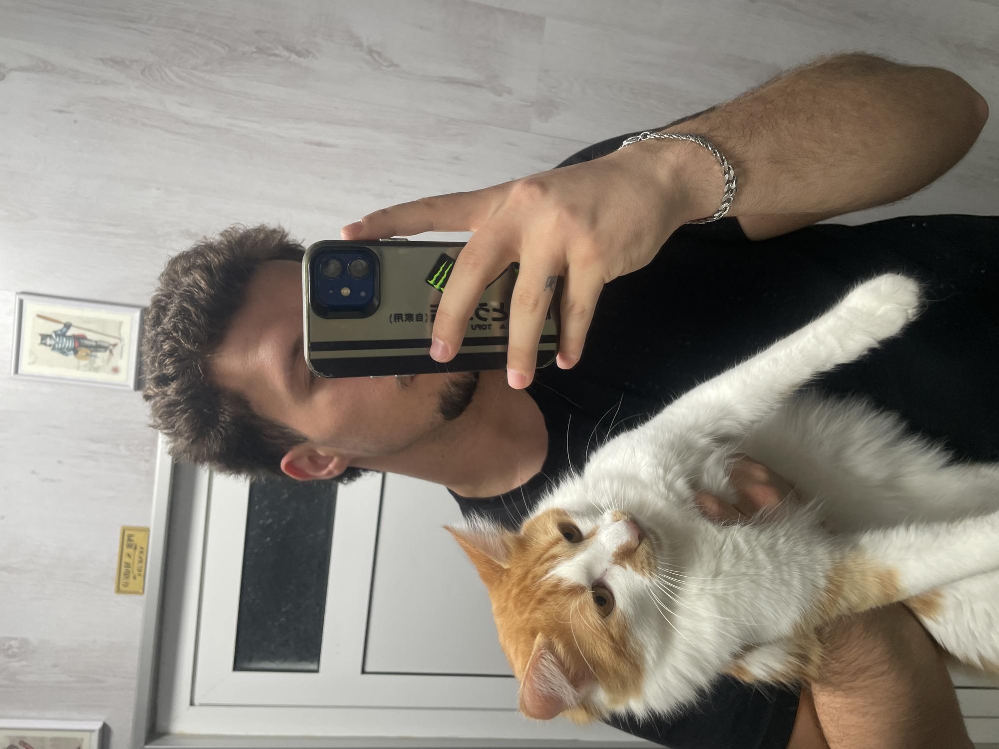 A man in a black shirt is holding a large orange and white cat, taking a selfie in what appears to be an indoor environment. He is holding up a smartphone with a distinctive case, which has stickers on it. The man has curly brown hair, a beard, and is wearing a silver bracelet on his left wrist. His hand also has a tattoo. The cat is looking at the camera. Behind the man, there is a white wall, a door, and a picture frame. There is also a small gold plaque. There is no indication of the city of Razgrad, Bulgaria in the image.