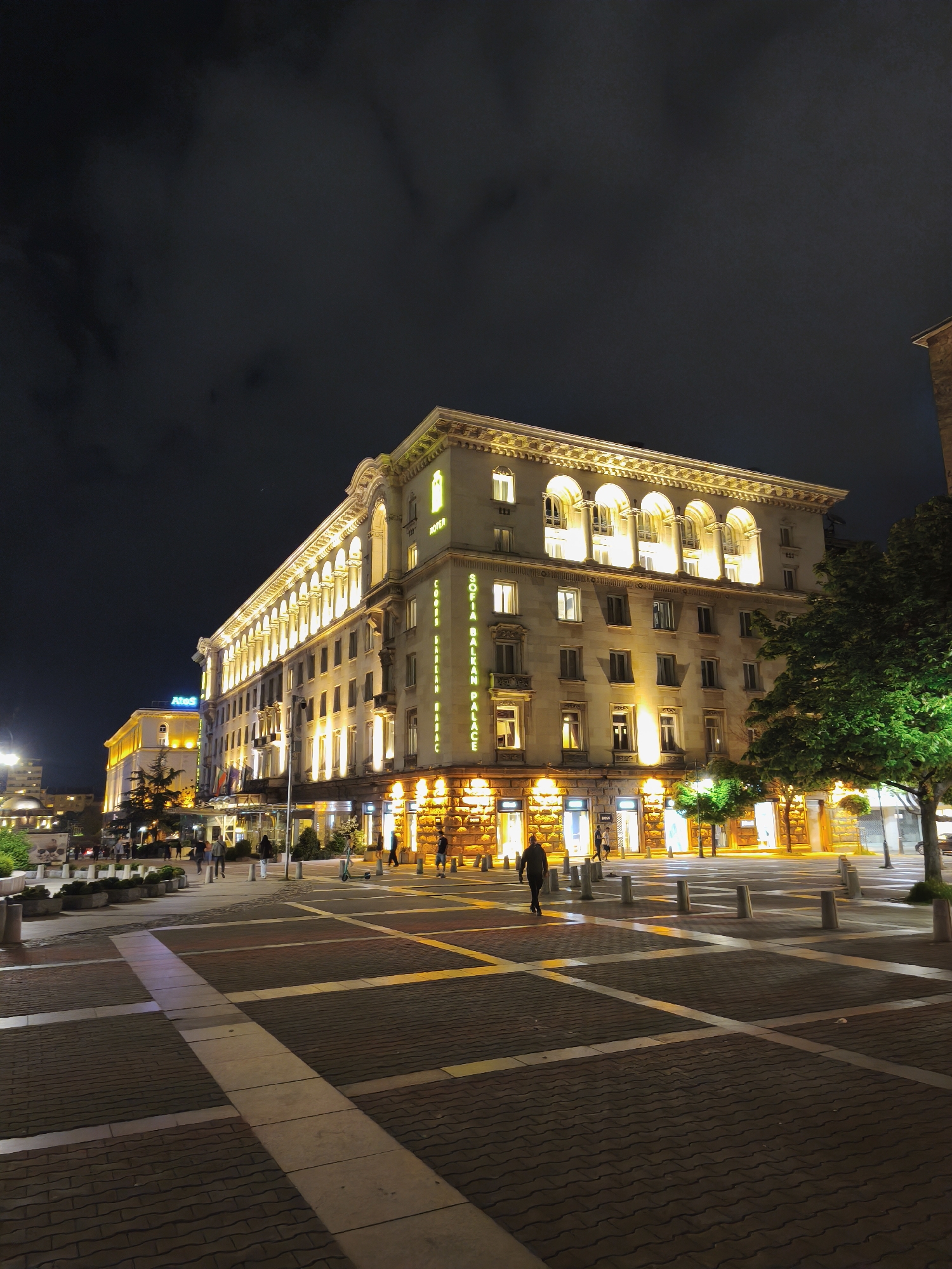 The image depicts a nighttime scene in Sofia, Bulgaria, featuring the Sofia Balkan Palace Hotel. The hotel, a large, multi-story building with illuminated windows and architectural detailing, dominates the frame. The area in front of the hotel is a paved plaza with a grid pattern and bollards. Pedestrians are walking across the plaza. The sky is dark, with some cloud cover. Other buildings are visible in the background, also illuminated at night. A tree with green leaves is on the right side of the image.