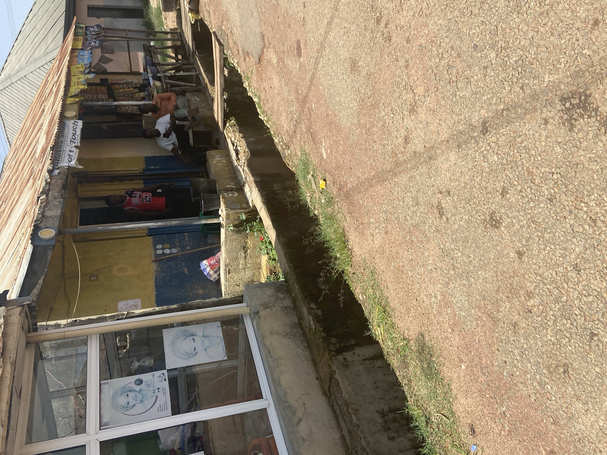 The image depicts a scene on a dusty street in Jos, Nigeria, outside a small shop. The shop has a corrugated metal roof, a yellow and blue painted facade, and a glass storefront. Inside the shop, a young man wearing a red and black basketball jersey with "BULLS 23" is standing near the entrance. Two other men are seated on a bench outside the shop, one looking at his phone and the other in an orange shirt. In front of the shop, a concrete drain runs along the edge of the street, covered in places by wooden planks. The ground is a mix of reddish-brown dirt and small stones. Various items are visible, including posters on the shop window, bags inside the shop, and products displayed on shelves outside.