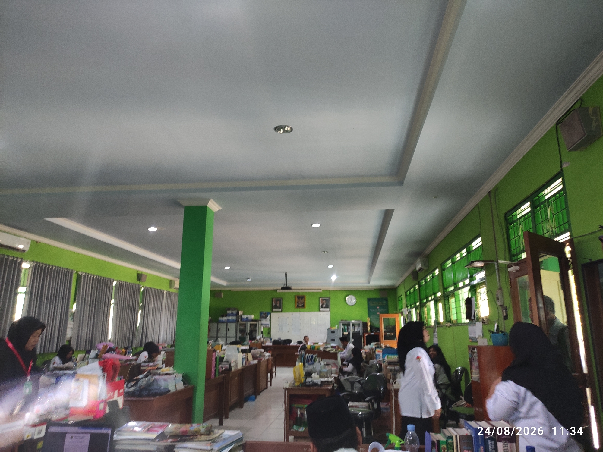 This image depicts an indoor scene, likely an office or classroom, with a green-painted wall on the right side and a lighter green wall on the left. The ceiling is white with visible light fixtures and a decorative molding. Several individuals are present, seated at desks and engaged in various activities, suggesting a working or studying environment.
On the left side of the frame, a person wearing a black hijab and a red lanyard is visible, looking down at a desk. Further back, another person in a black hijab is also seated at a desk. The desks appear to be made of wood and are cluttered with papers and other items.
In the central part of the image, more people are seated at desks. One person is wearing a white shirt and a black hijab. Another person is seated behind a desk, seemingly interacting with a computer. A projector is mounted on the ceiling.
On the right side of the image, there are bookshelves and what appears to be a cabinet. A tall, framed mirror is leaning against the wall. A person in a white shirt and a black hijab is partially visible, facing away from the camera. Another person in a black hijab is also visible.
The environment appears to be well-lit, with natural light coming from windows covered by grey curtains on the left. The overall impression is of a busy, functional indoor space. The timestamp "24/08/2026 11:34" is overlaid on the bottom right corner of the image. There is no direct indication of the location being Tamanrejo, Indonesia within the visual data.