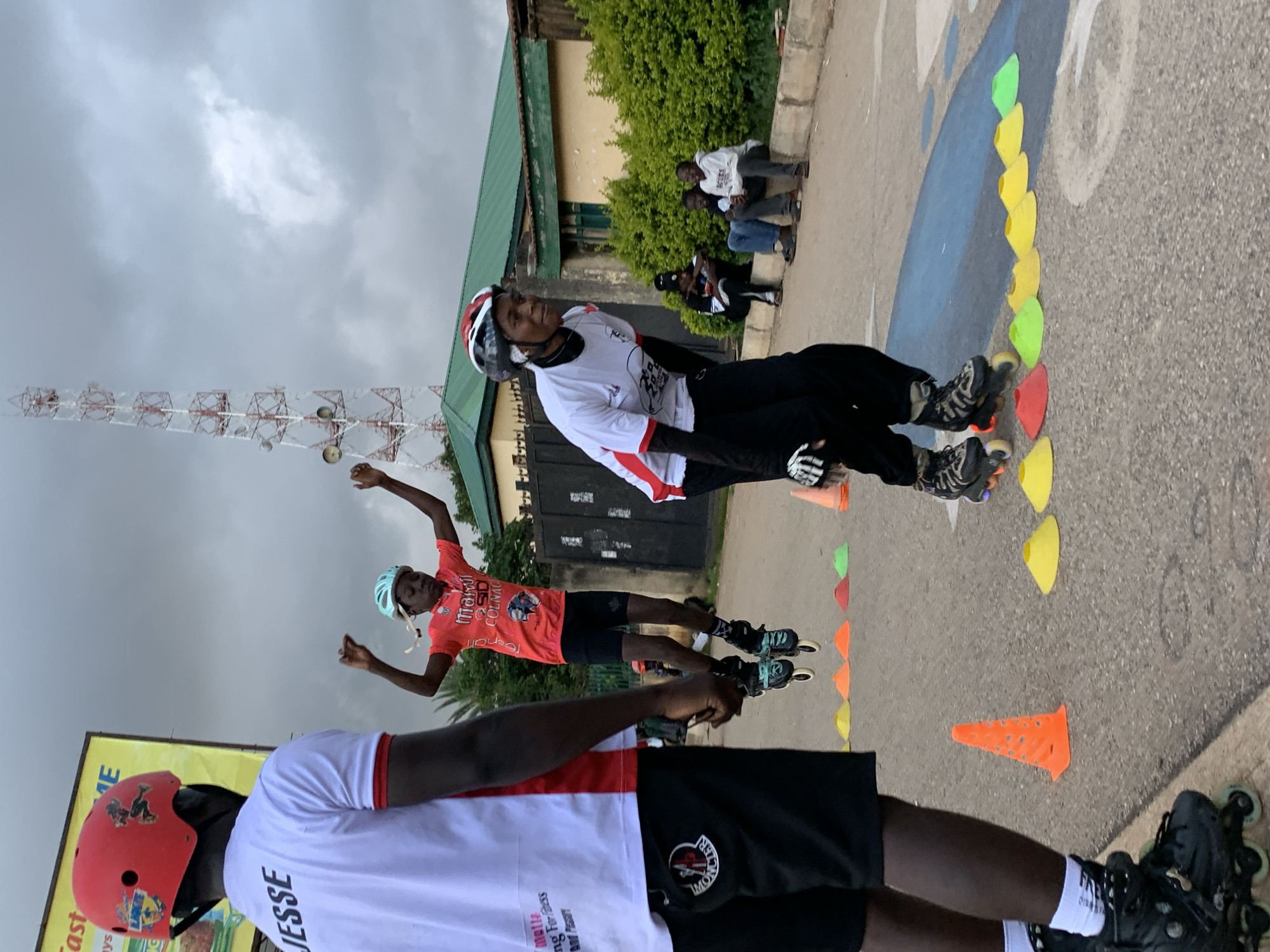 In Jos, Nigeria, under an overcast sky, three individuals engage in inline skating. A person in the foreground, viewed from the waist down, wears black inline skates, white socks, and black shorts with a visible Moncler logo. To their left, a person in a red helmet and white shirt with "JESSE" printed on it is partially visible. In the middle ground, a young person in a light blue helmet and an orange shirt leaps mid-air, wearing black inline skates. Another individual, to the right, wearing a red and white helmet, white shirt, and black pants, is also inline skating. A line of colorful cones, including orange, yellow, red, and green, are set up on the asphalt, suggesting a training or practice area. In the background, a tall communication tower rises against the sky, and a building with a green roof is visible, along with some seated individuals. The overall scene depicts an outdoor sports activity in an urban environment.